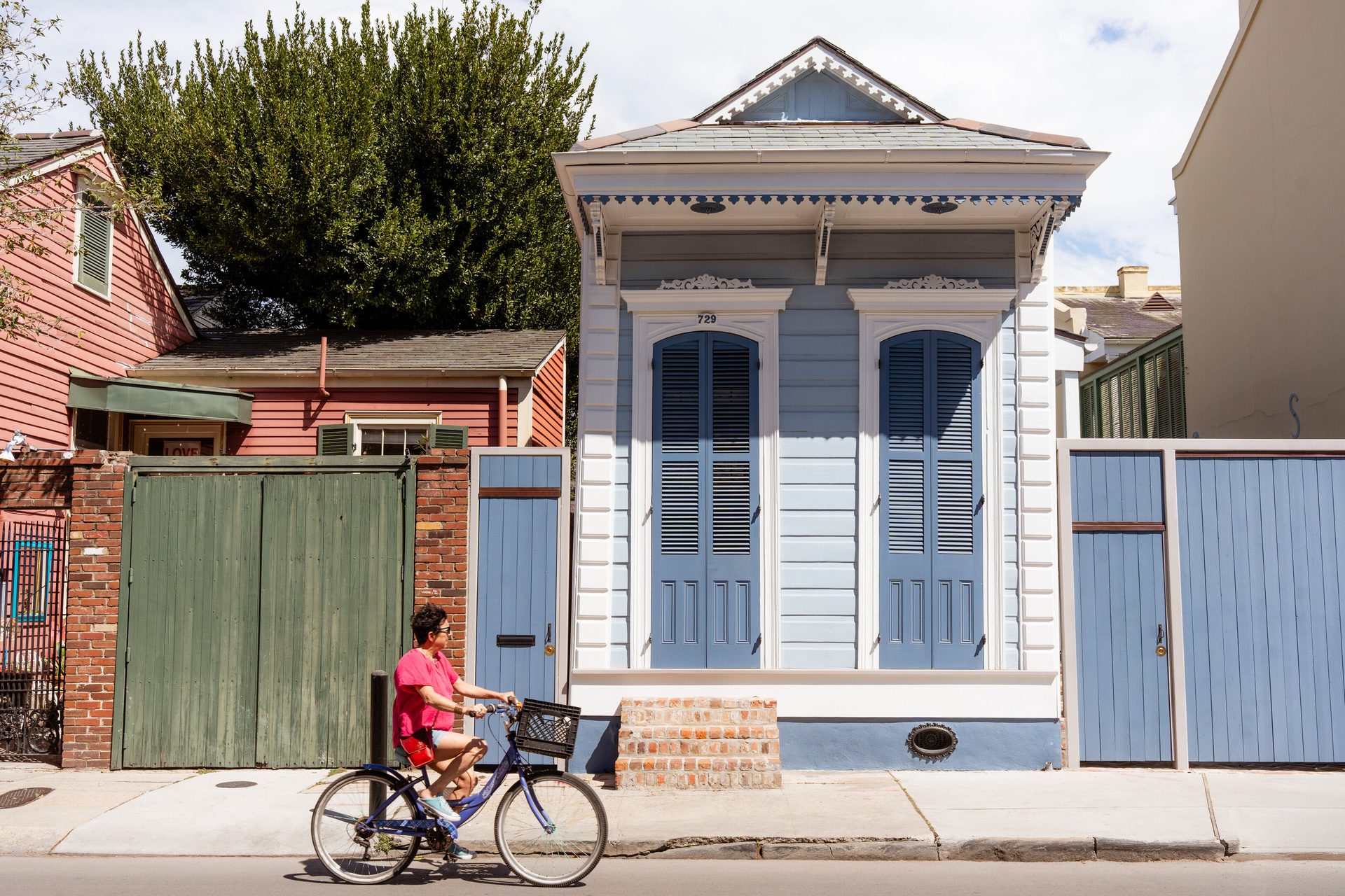 A woman rides a bicycle past colorful houses, including a light blue shotgun house and a red house with a green gate.