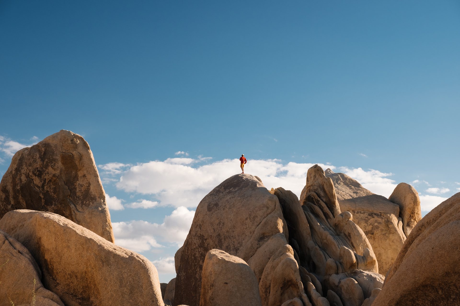 A person in red atop a huge desert rock formation under a blue, cloudy sky.