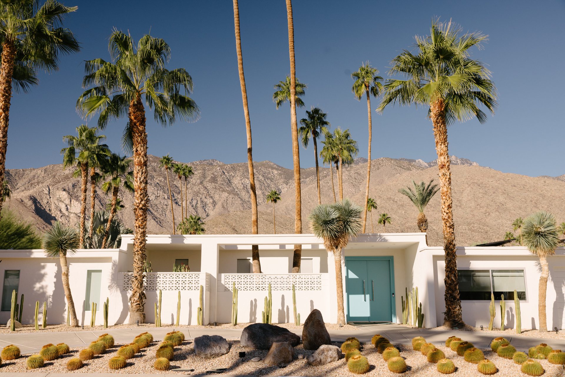 White house with a teal door, palm trees, cacti, and desert mountains under a clear sky.