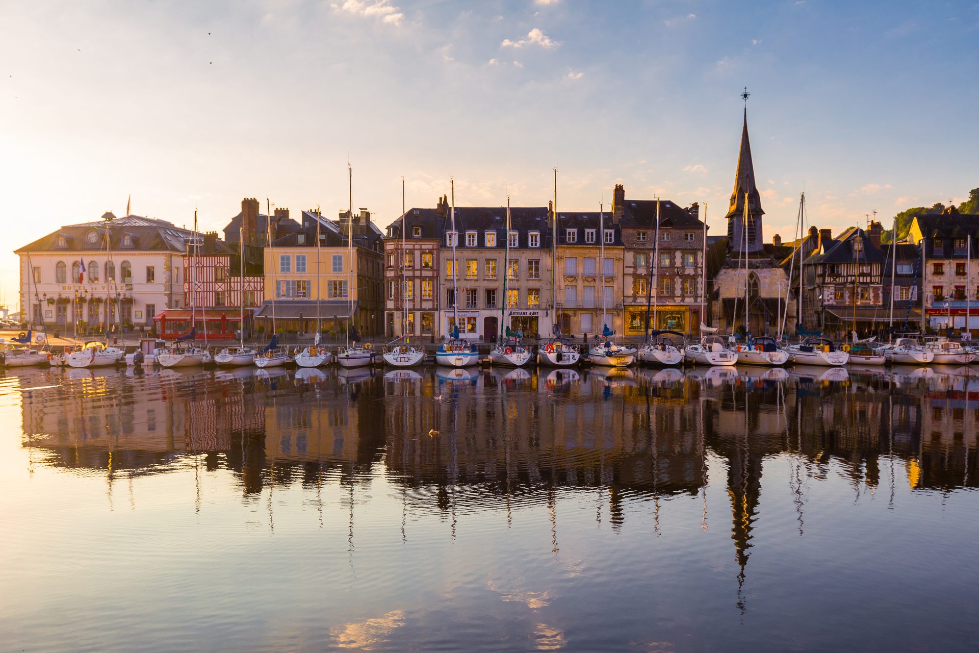 Historic harbor with colorful buildings, sailboats, and their reflections at sunrise.