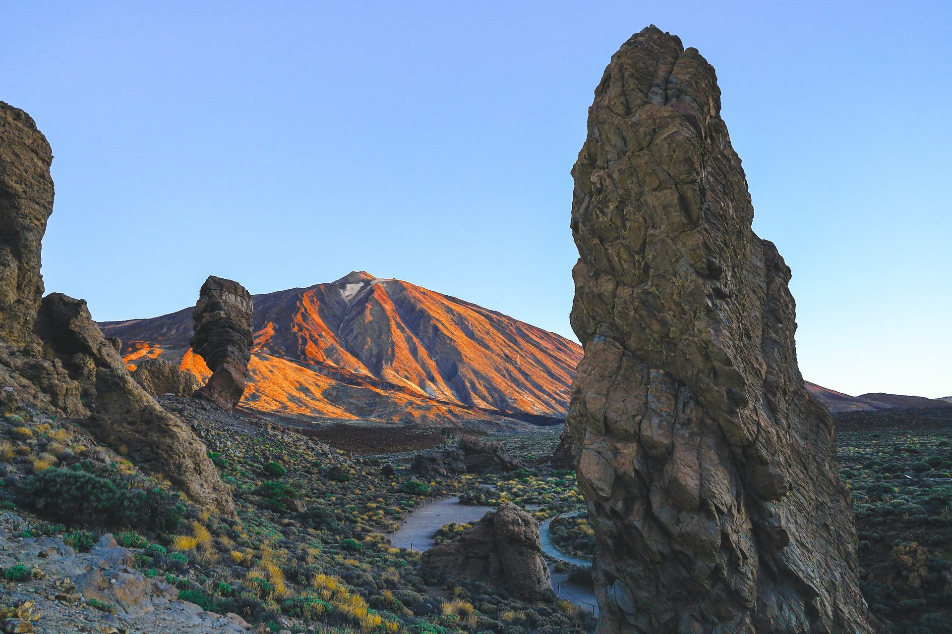 Teide volcano bathed in golden light at sunset, framed by a tall rock spire and other formations.