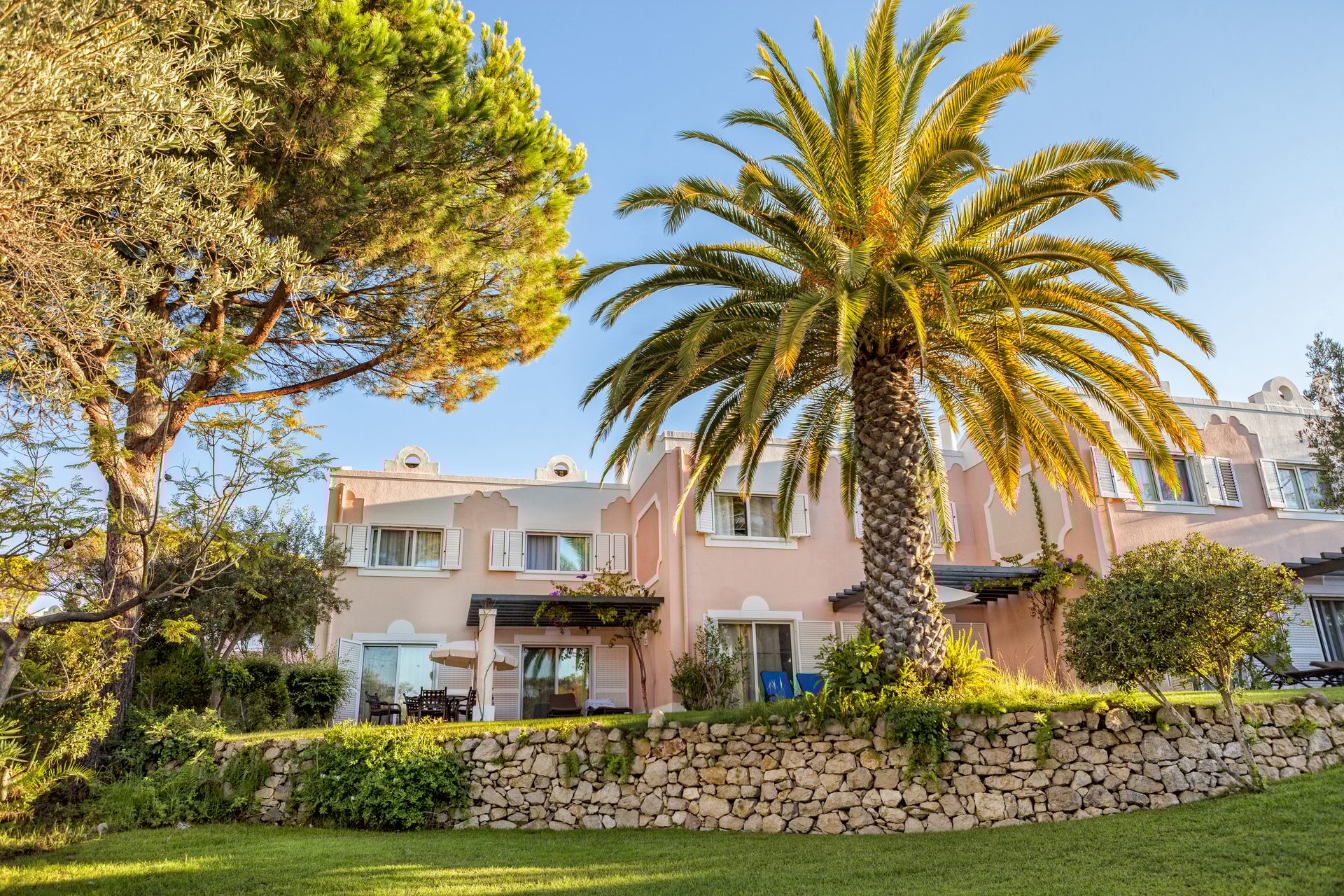 Pink hotel building with palm and other trees, stone wall, and green grass under a clear sky.
