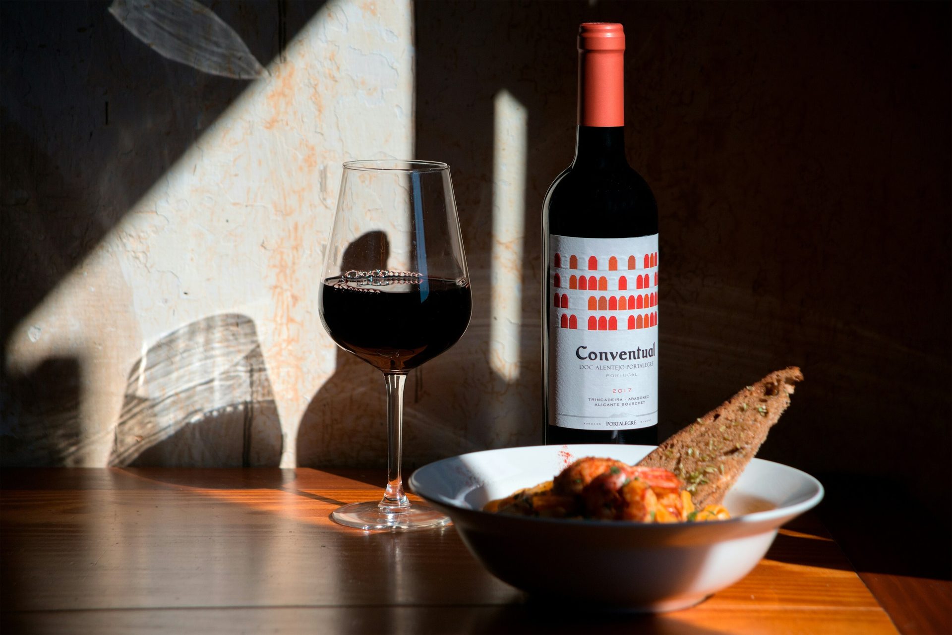 Red wine in glass & 'Conventual' bottle, food with toast, on a sunlit wooden table.