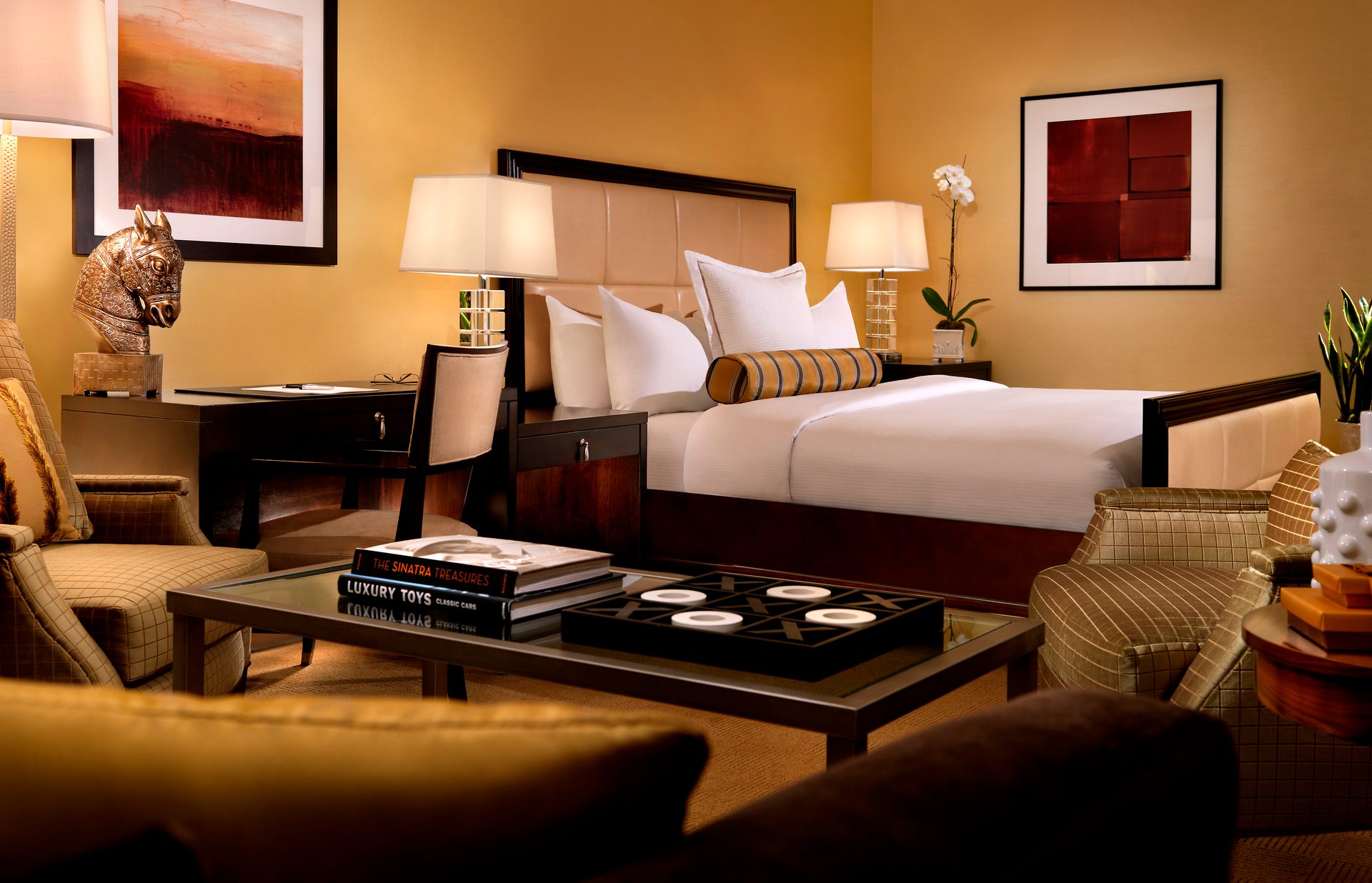 Luxurious hotel room with a bed, desk, and sitting area, featuring warm golden tones.