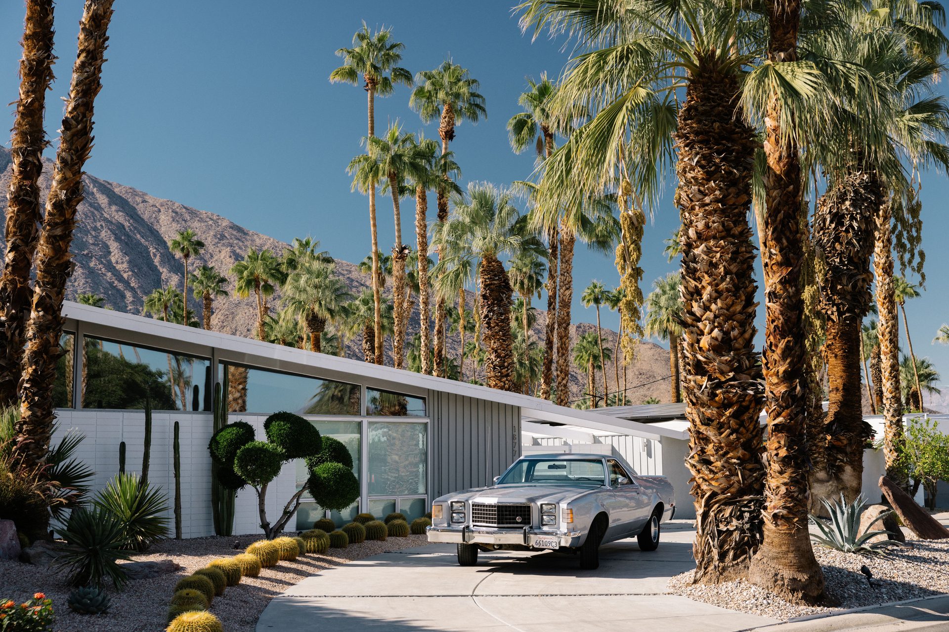 Vintage silver car at a modern desert house with palm trees and mountains.