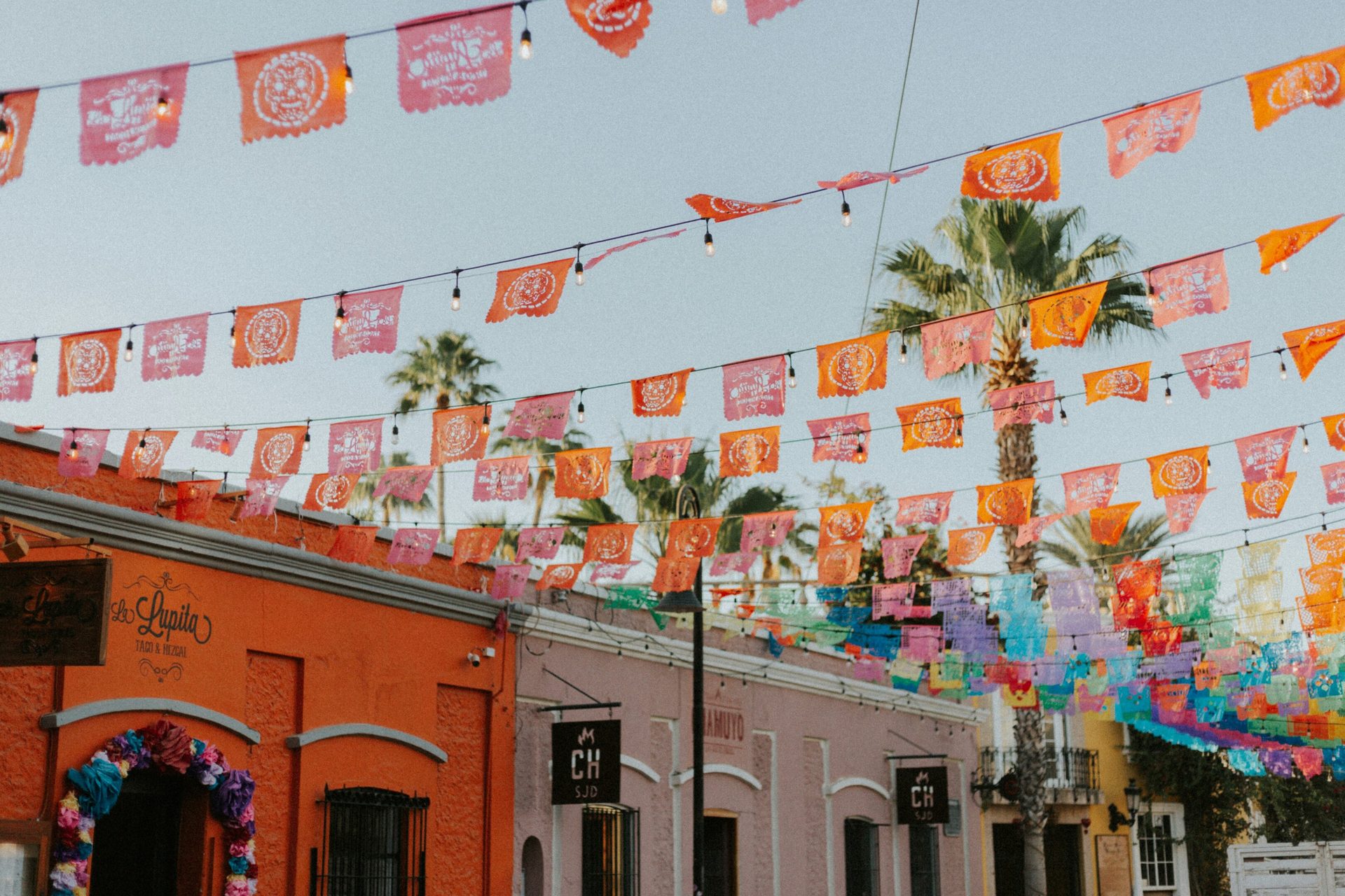 Papel picado flags strung over a street with colorful buildings and palm trees.