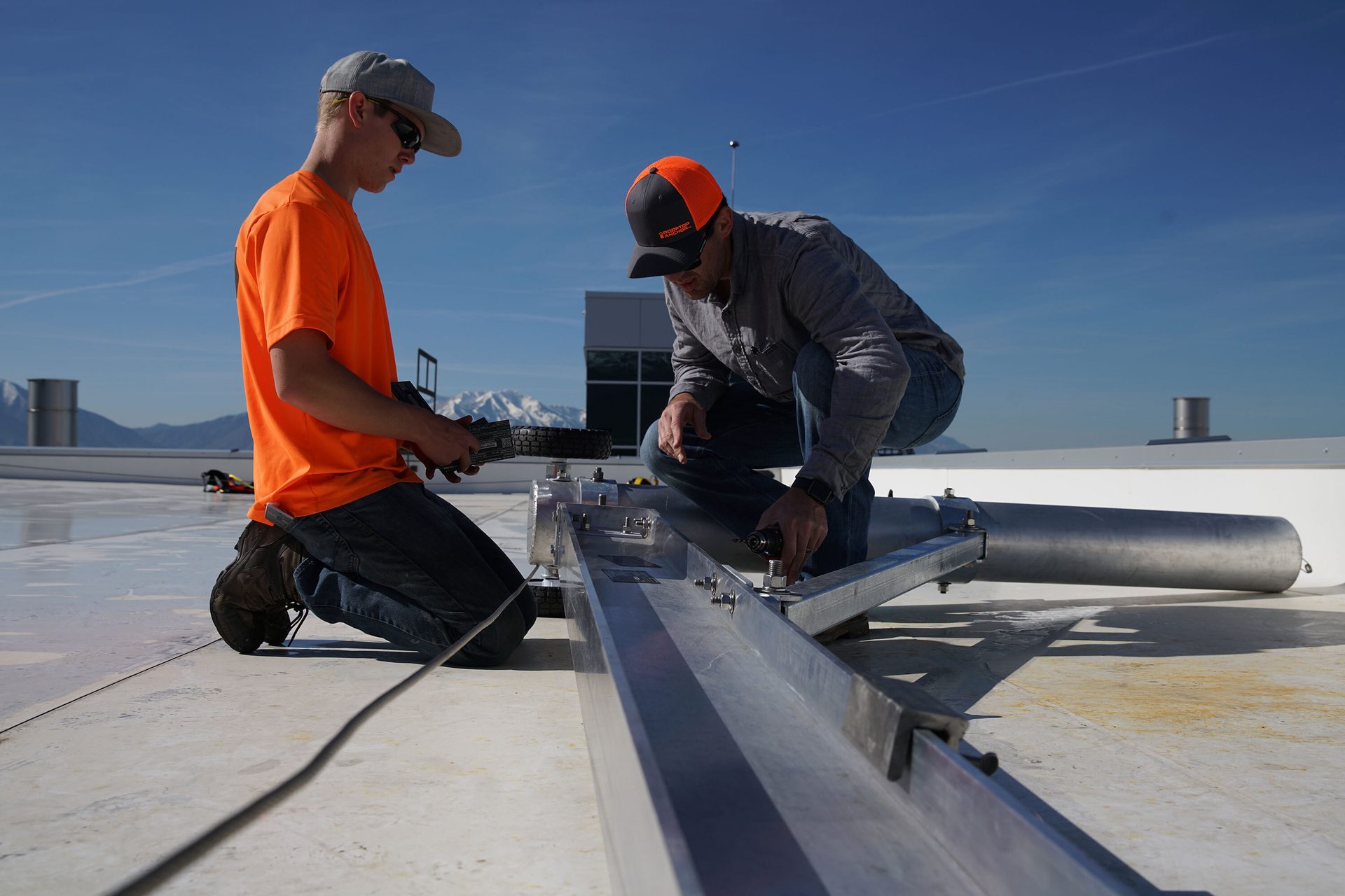 Two men on a rooftop assembling a metal structure, with mountains in the background.