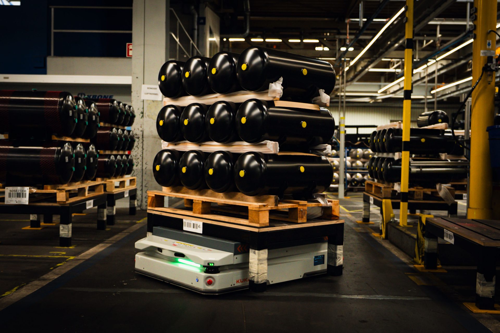 An AGV transports black industrial tanks stacked on pallets in a factory.