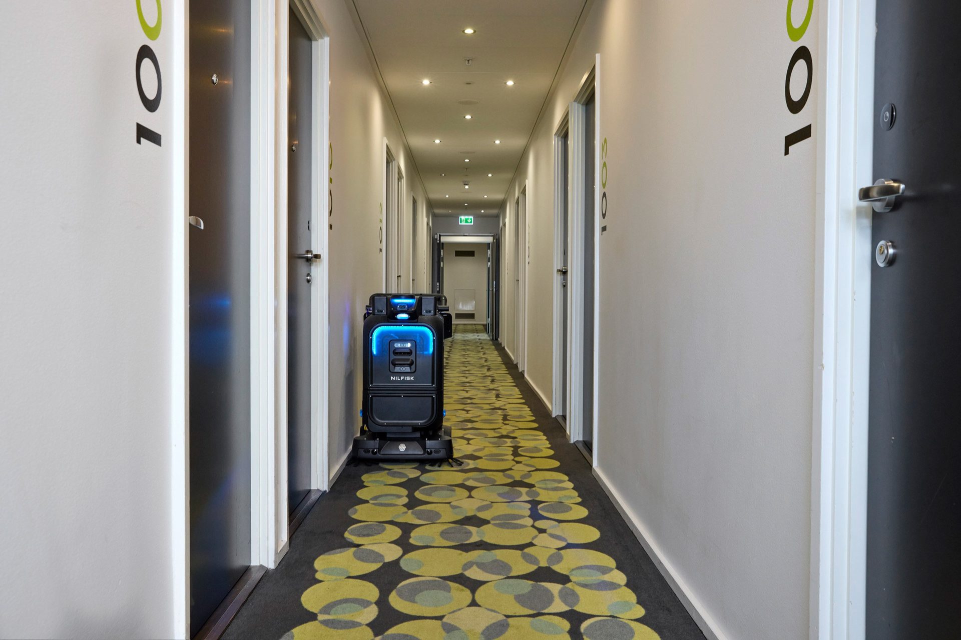 A Nilfisk cleaning robot in a hotel hallway with patterned carpet.