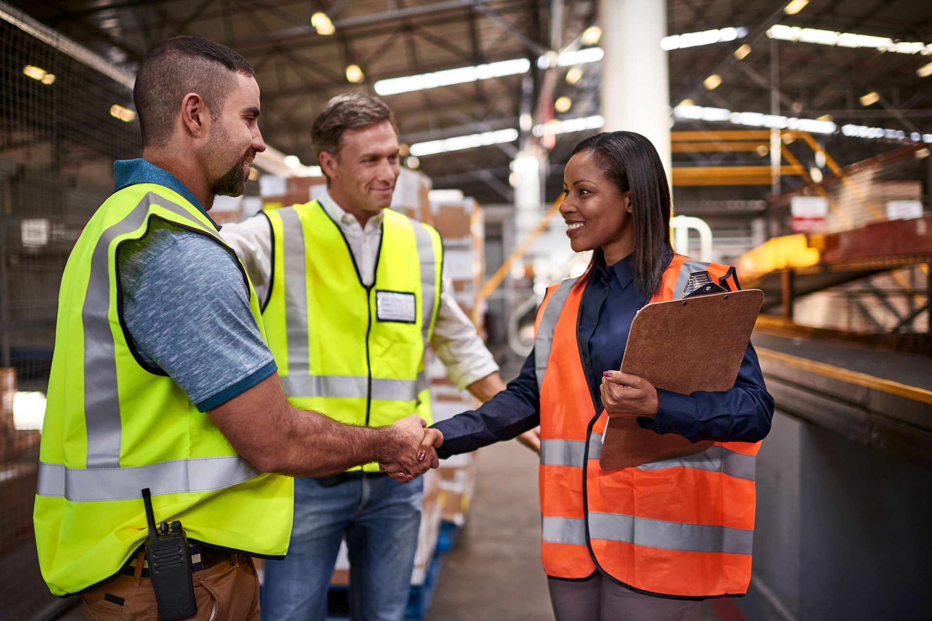 Three warehouse workers, two men and one woman, shaking hands and smiling in a warehouse.