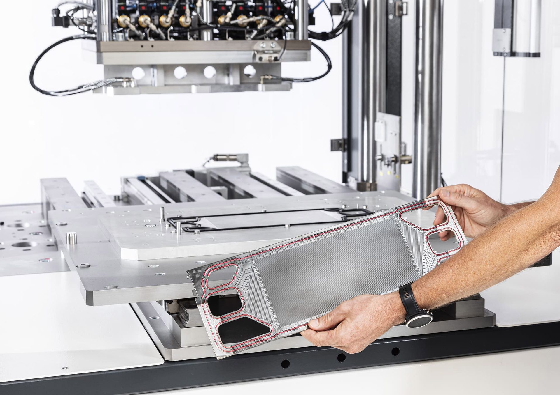 Hands holding a metal heat exchanger plate with a red gasket over industrial machinery.