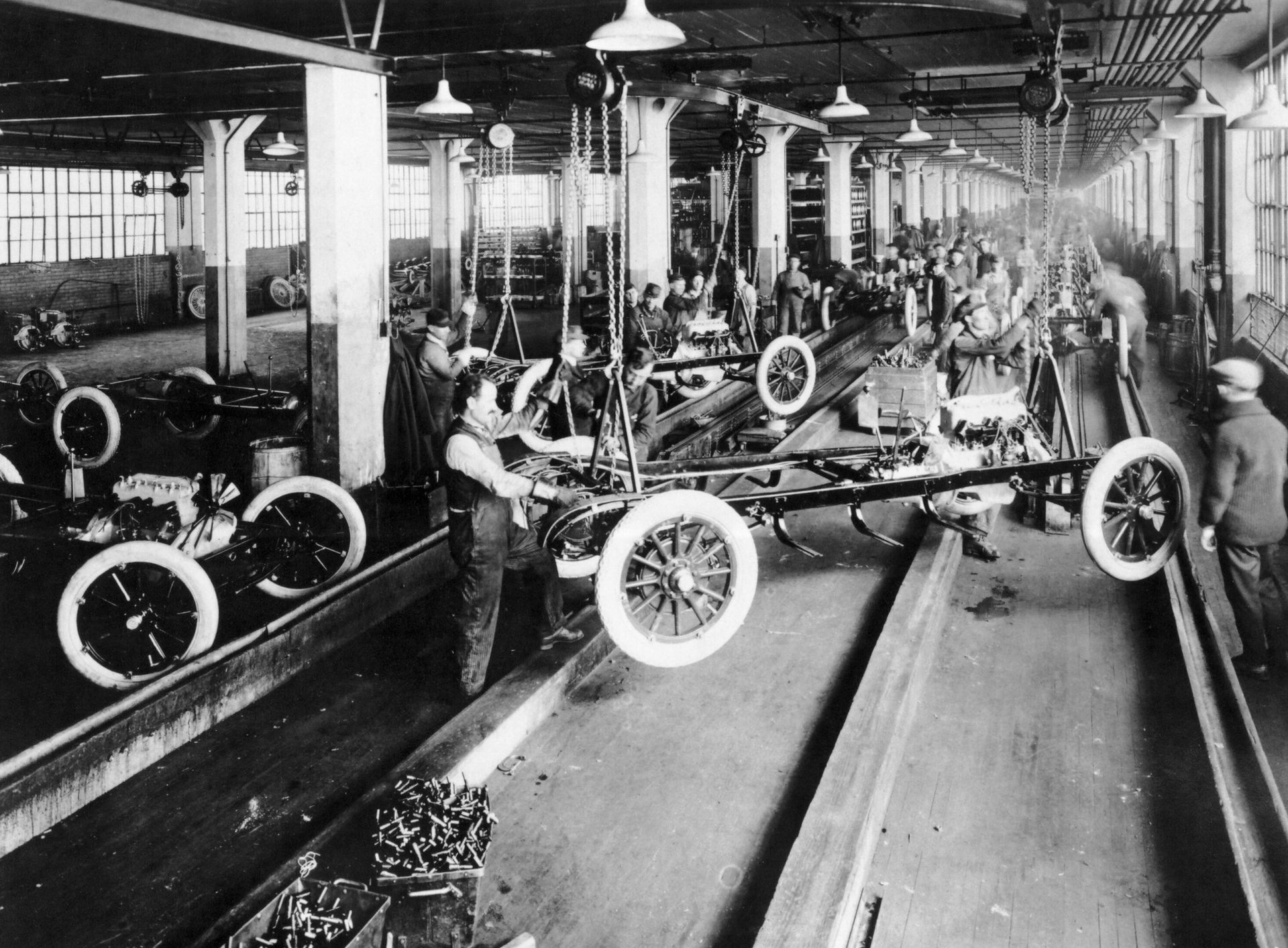 Early 20th-century factory assembly line with workers building car chassis and engines.