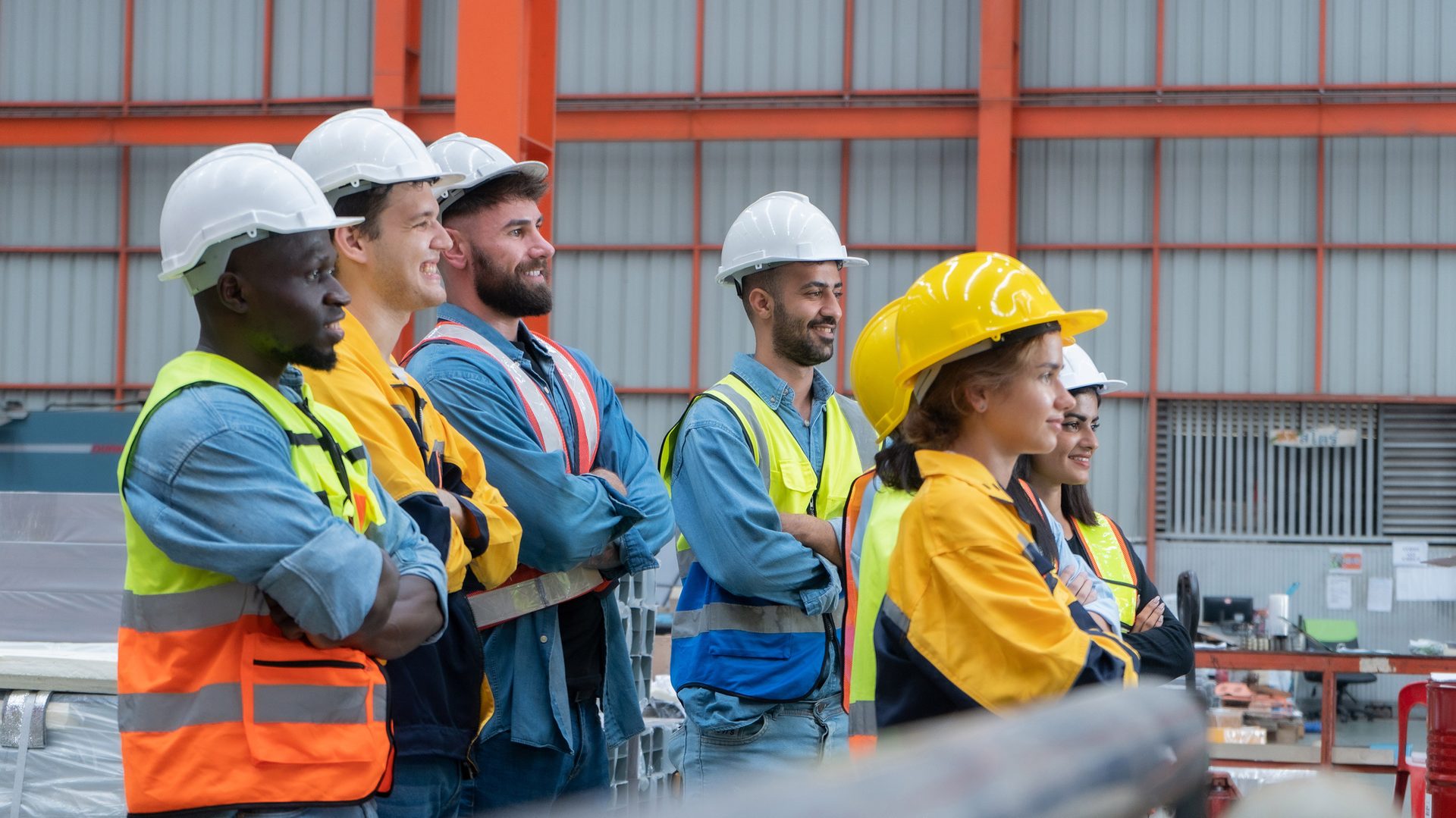 Smiling factory workers in hard hats and safety vests, looking right.