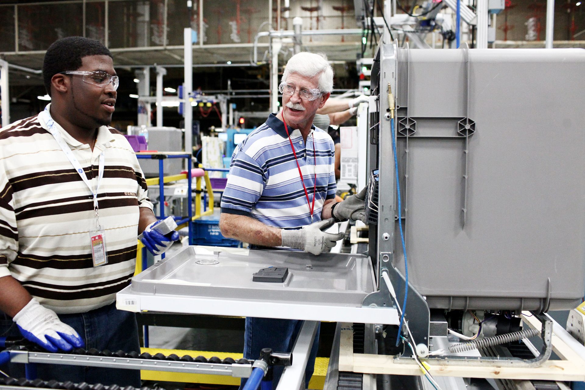 Two men, one Black, one white, assemble an appliance on a factory line.