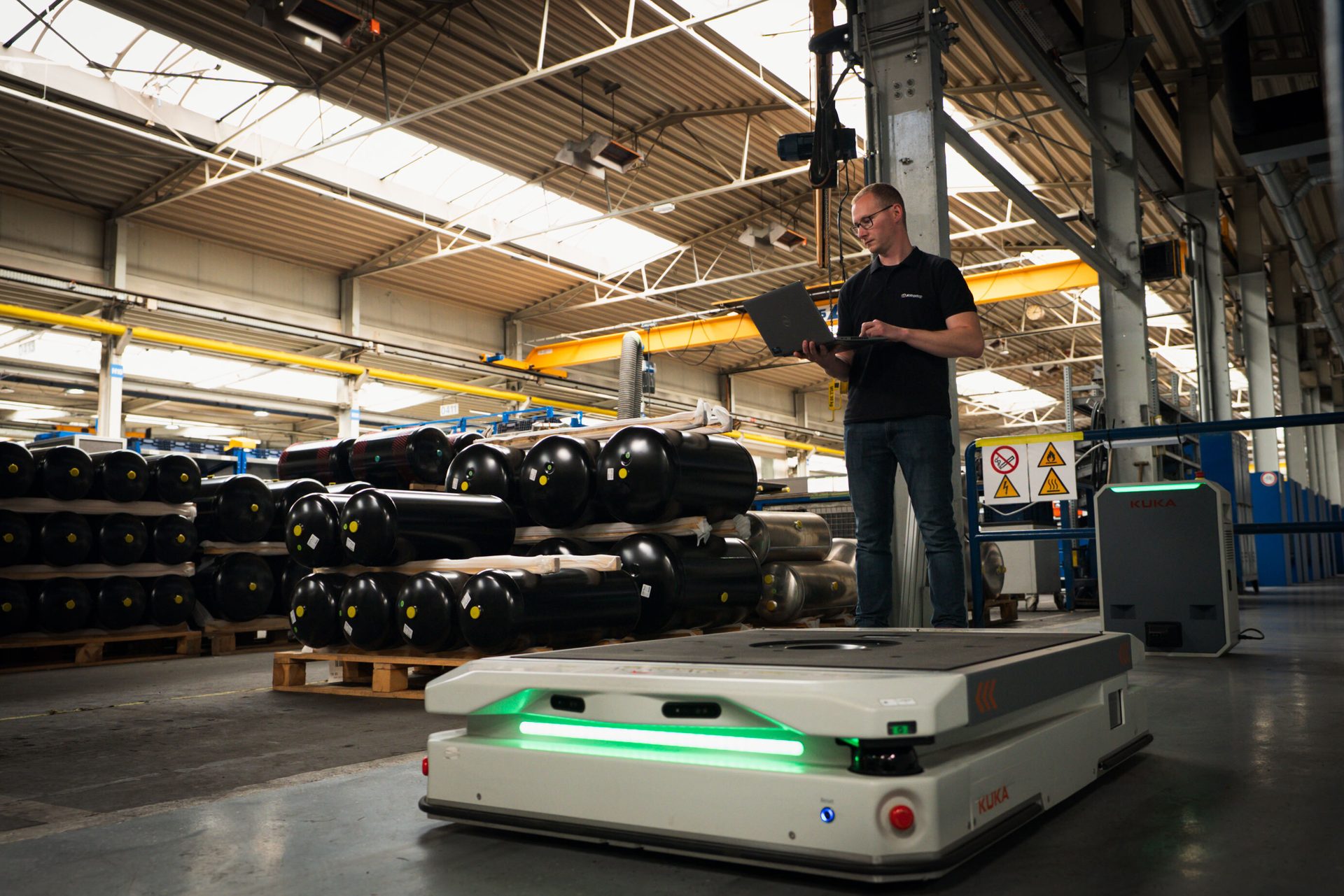 Man with laptop, Kuka autonomous robot, and stacked parts in a factory.