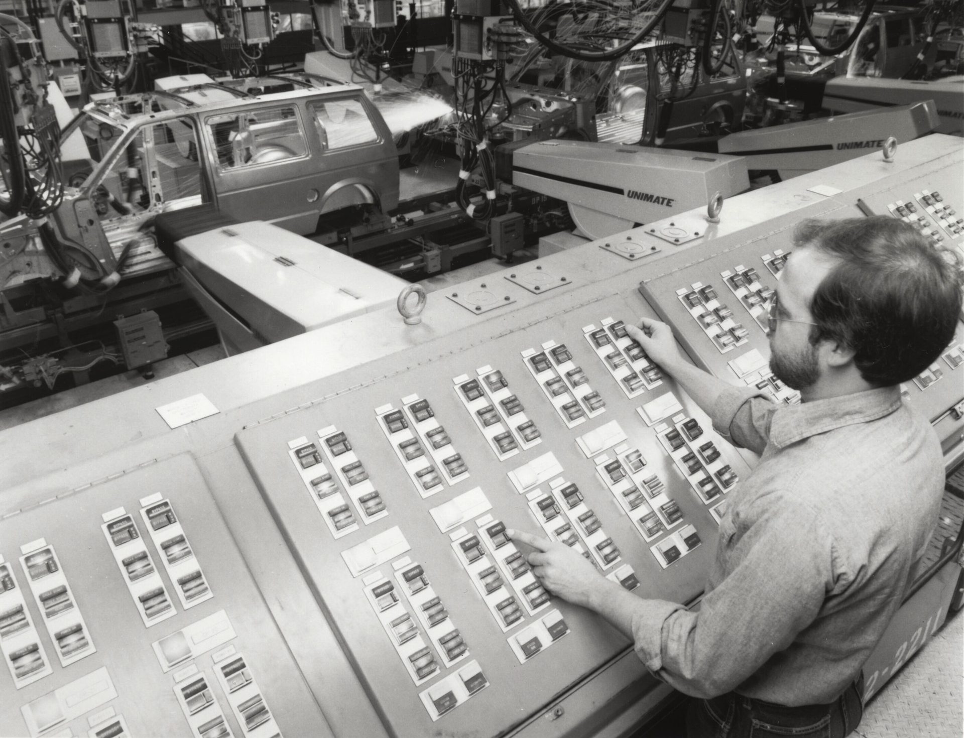 Man operates a control panel for car assembly robots (UNIMATE) in a factory.
