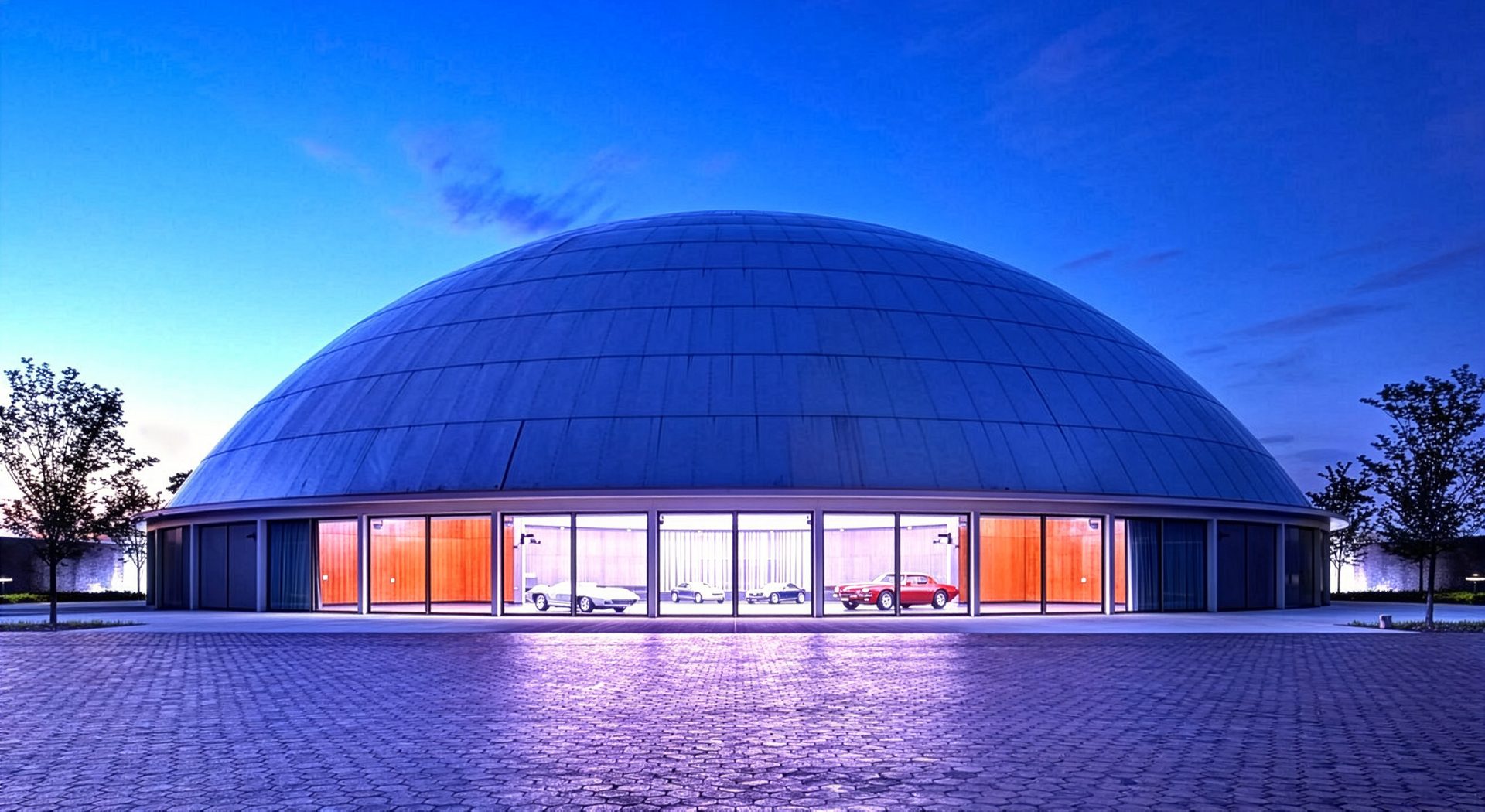 A large blue dome building displaying classic cars through illuminated windows at dusk.