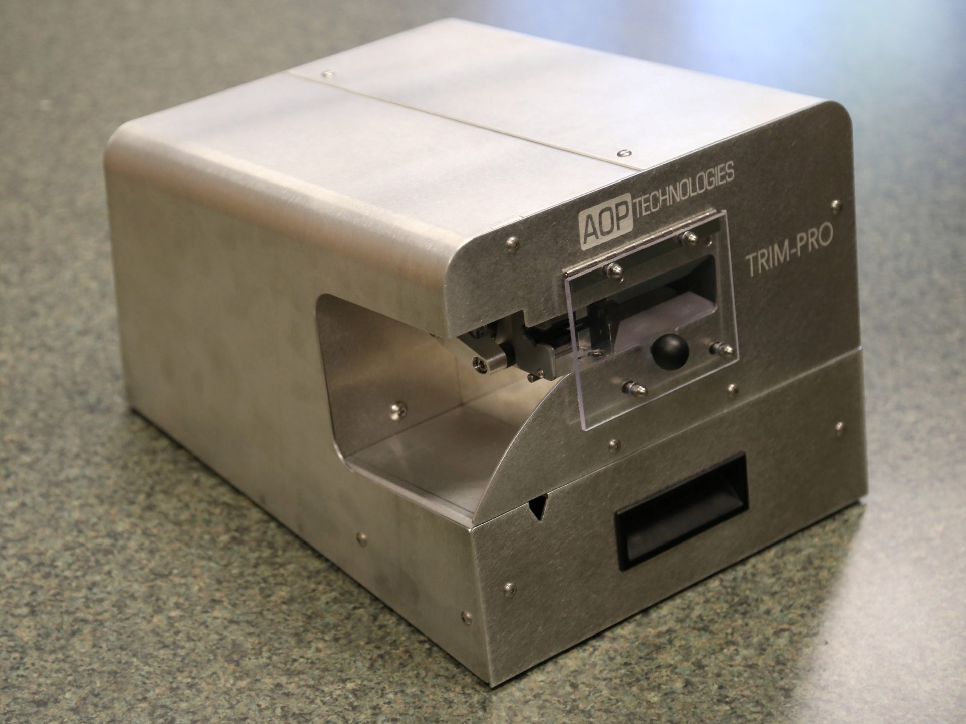 A silver metal AOP Technologies Trim-Pro machine, a compact industrial trimming device, on a speckled surface.