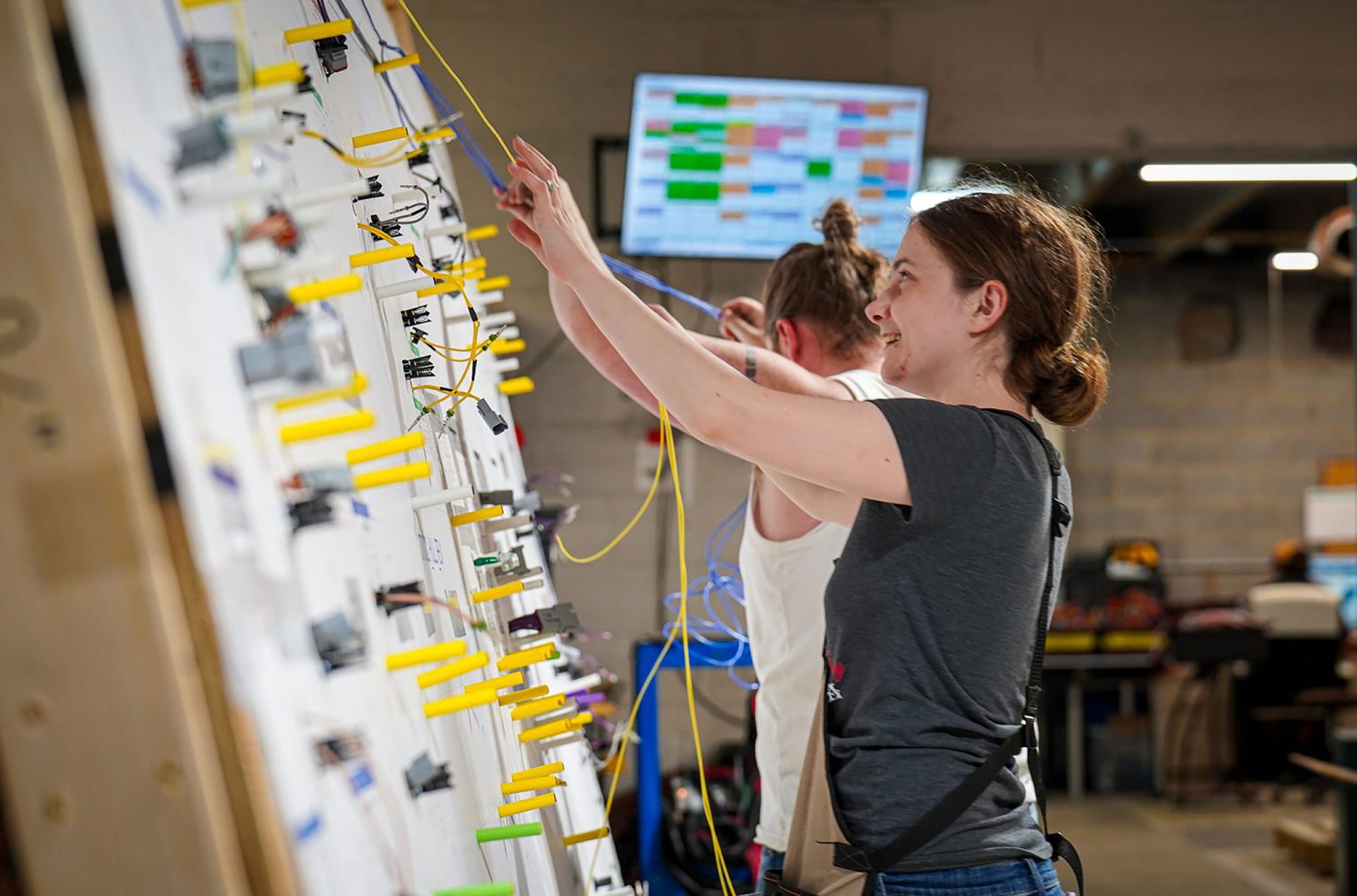Two women assemble electrical wiring harnesses on a large board in a workshop, smiling.