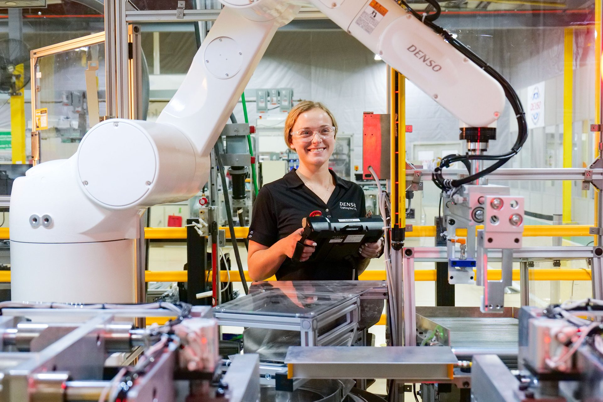 Smiling woman in safety glasses controls a white Denso robotic arm in a factory.