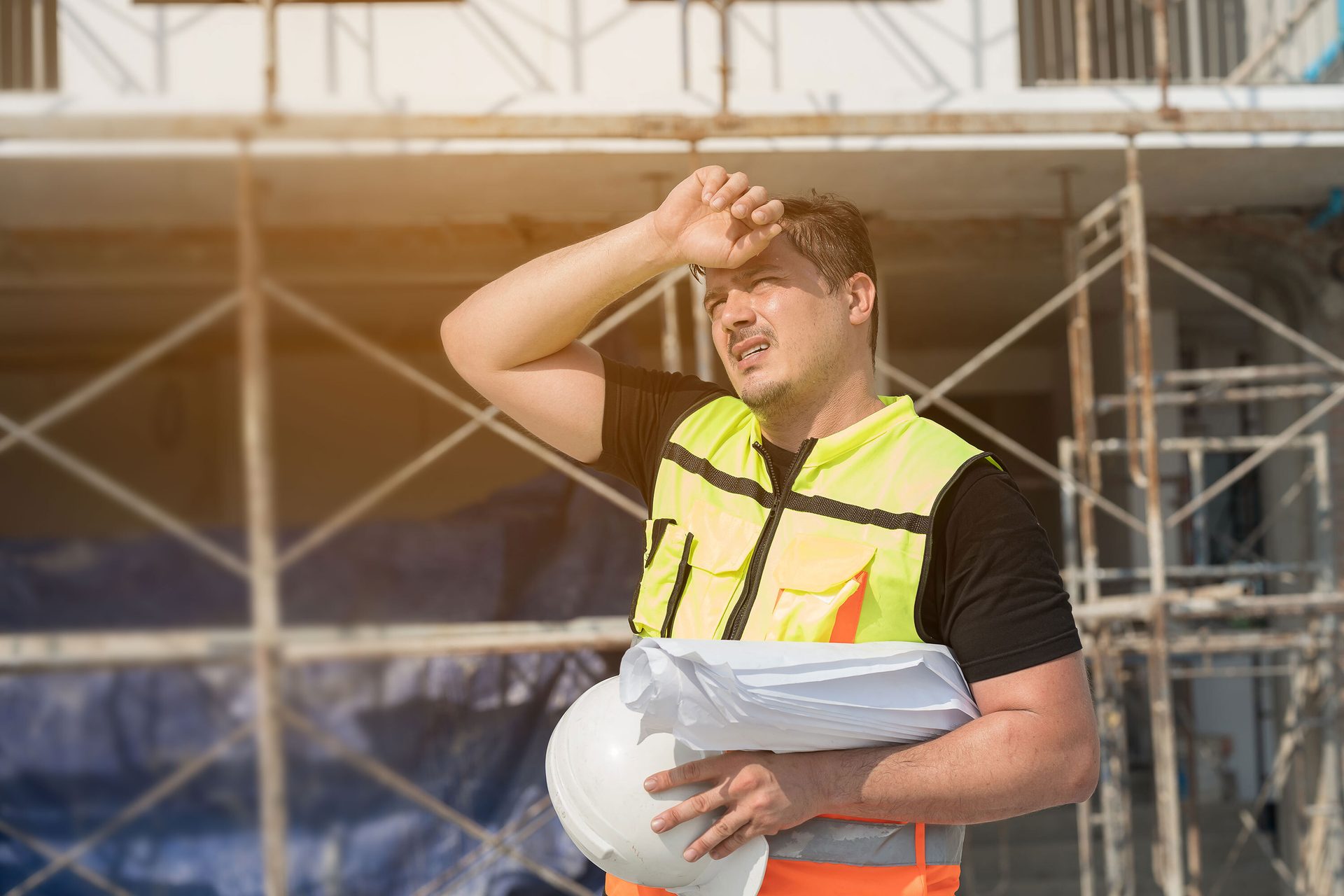 Man in safety vest sweating at construction site, holding hard hat and blueprints.