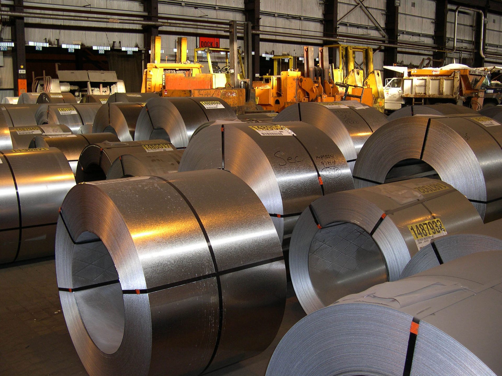 Many large rolls of coiled sheet metal in an industrial warehouse, with forklifts and trucks.