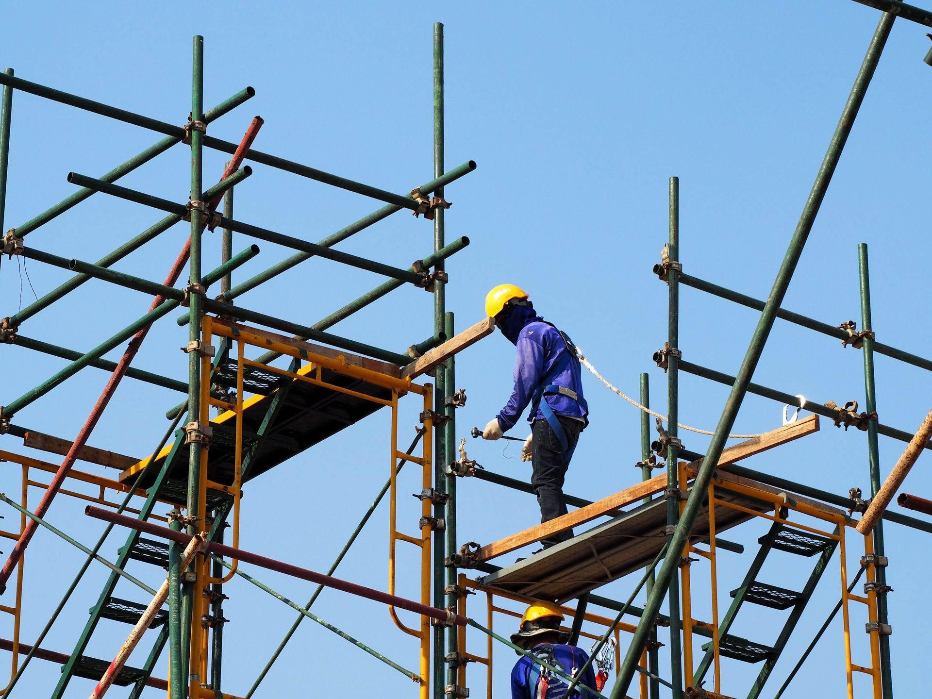 A construction worker in a hard hat and safety gear on scaffolding, assembling the structure against a blue sky.