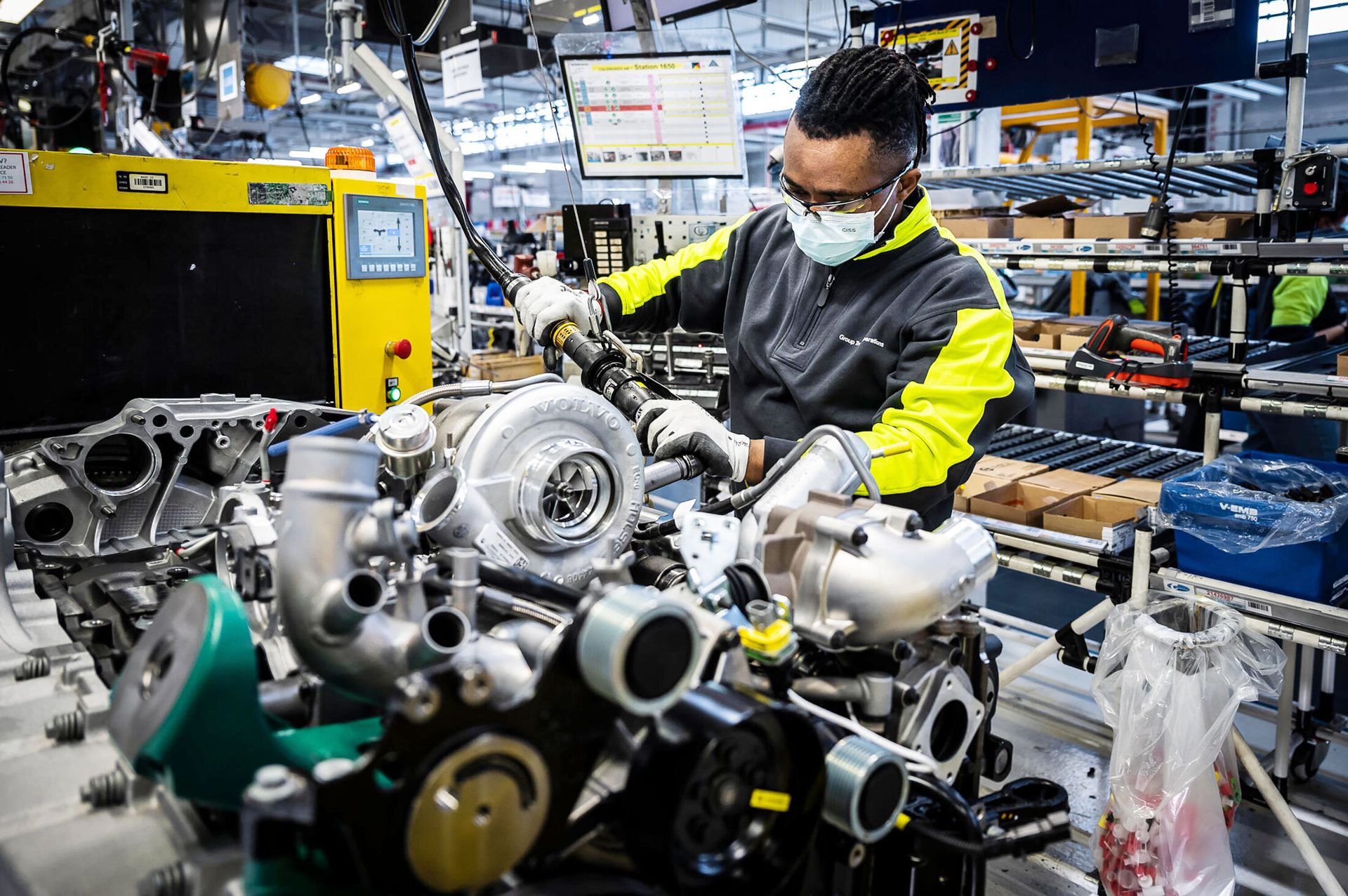 A worker wearing a mask and safety glasses assembles a Volvo engine on a production line.
