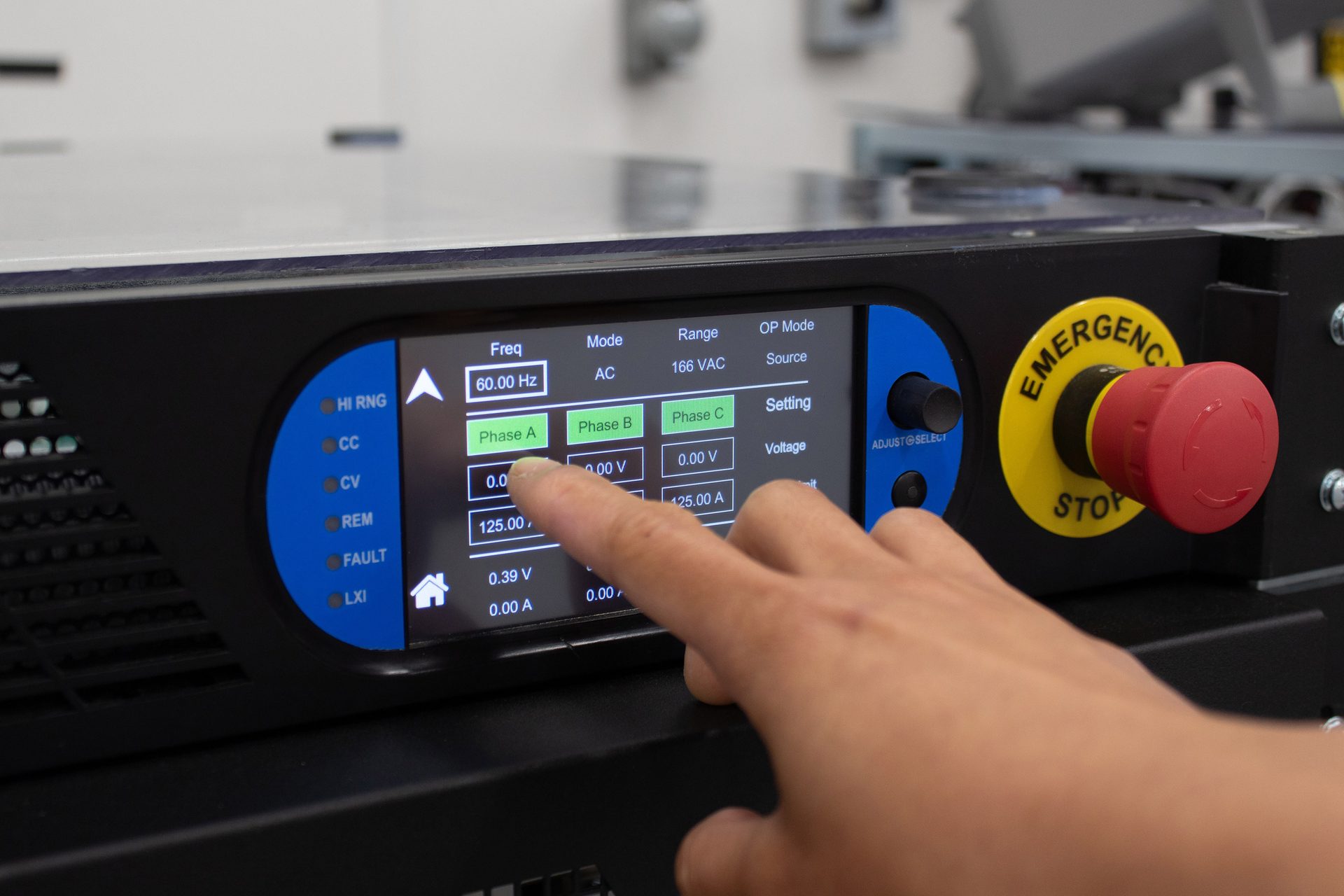 Hand interacting with a power supply's touchscreen showing electrical parameters and an emergency stop.