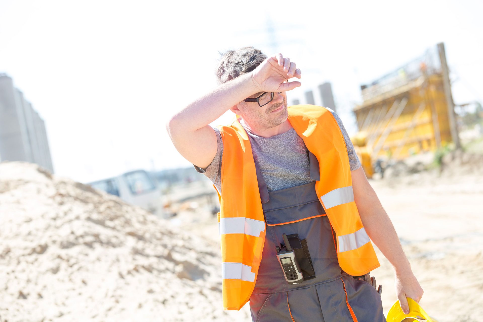 Construction worker shielding eyes from bright sun, holding hard hat.