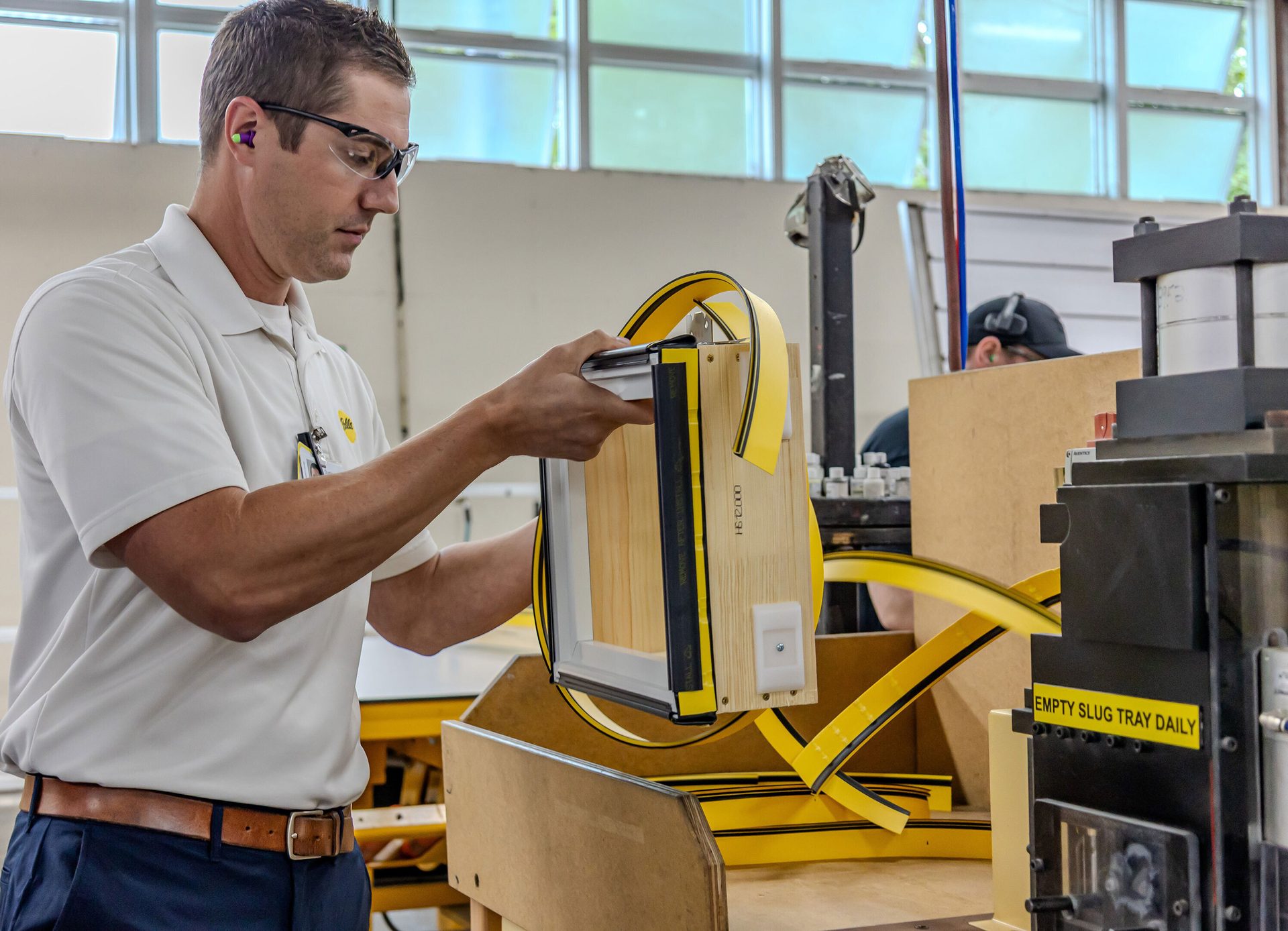 A worker in safety gear applies yellow banding to a window frame in a workshop.