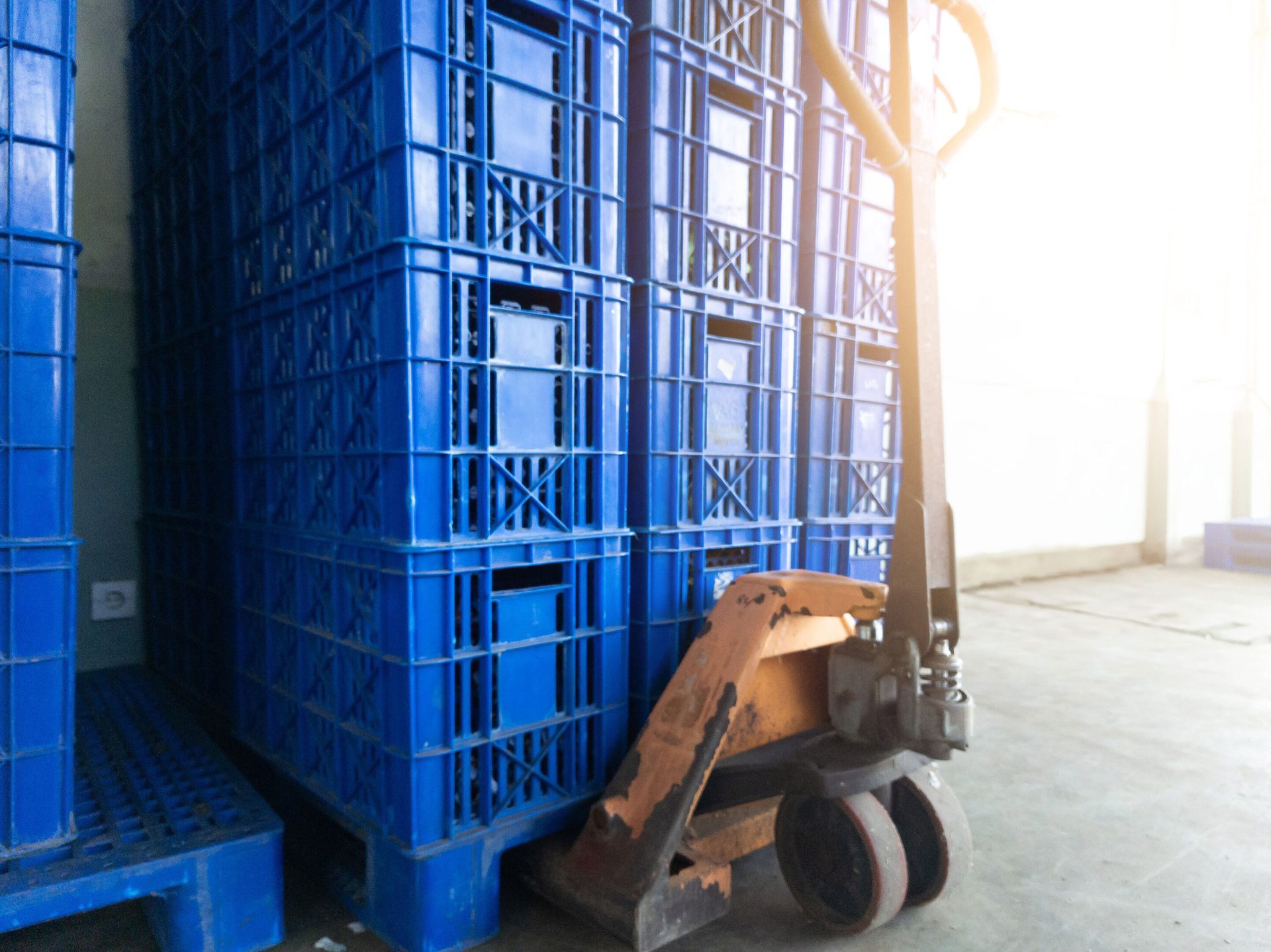 Stacks of blue plastic crates in a warehouse, with a rusted orange pallet jack in the foreground, illuminated by sunlight.