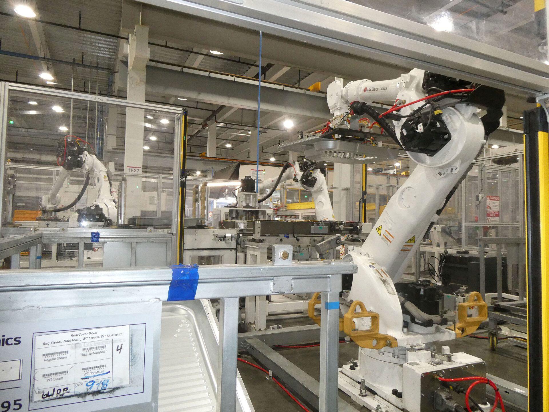 White LG Electronics robotic arms on an assembly line in a factory, a "RearCover Dryer" label visible.