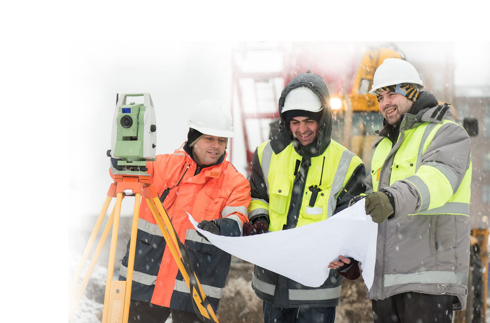 Three surveyors in winter gear review plans at a snowy construction site.