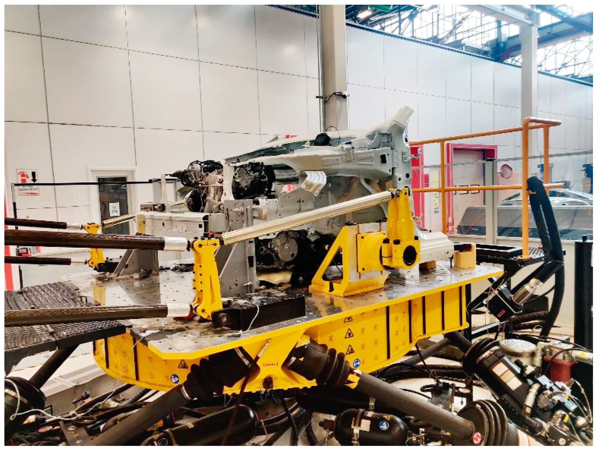 A gray car chassis is mounted on a yellow test rig with various hoses and equipment in an industrial setting.