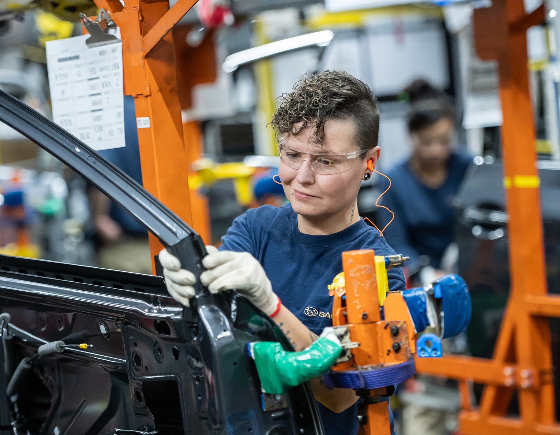 Woman with safety glasses and earplugs working on a car part assembly line in a factory.