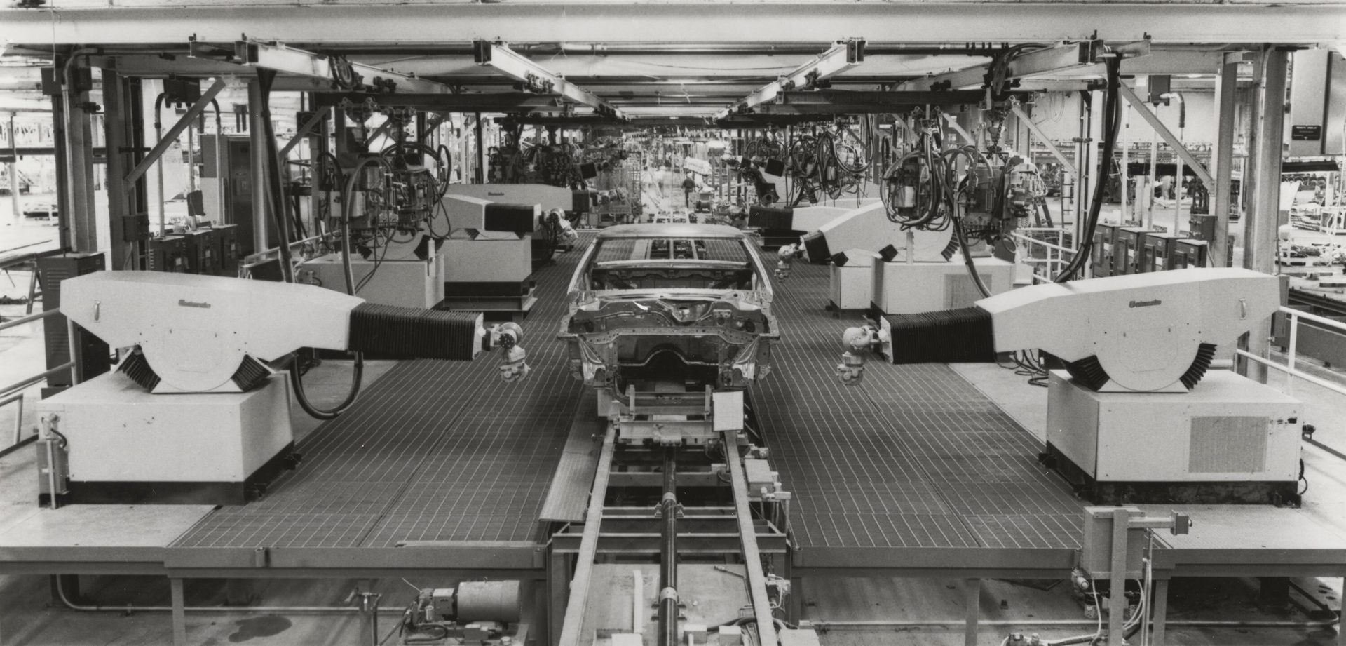 Black and white image of robotic arms working on a car body on an assembly line in a factory.