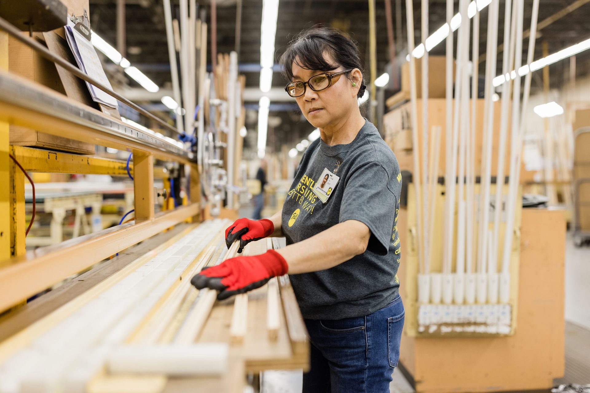 Asian woman in factory, glasses, gloves, handling wooden pieces.