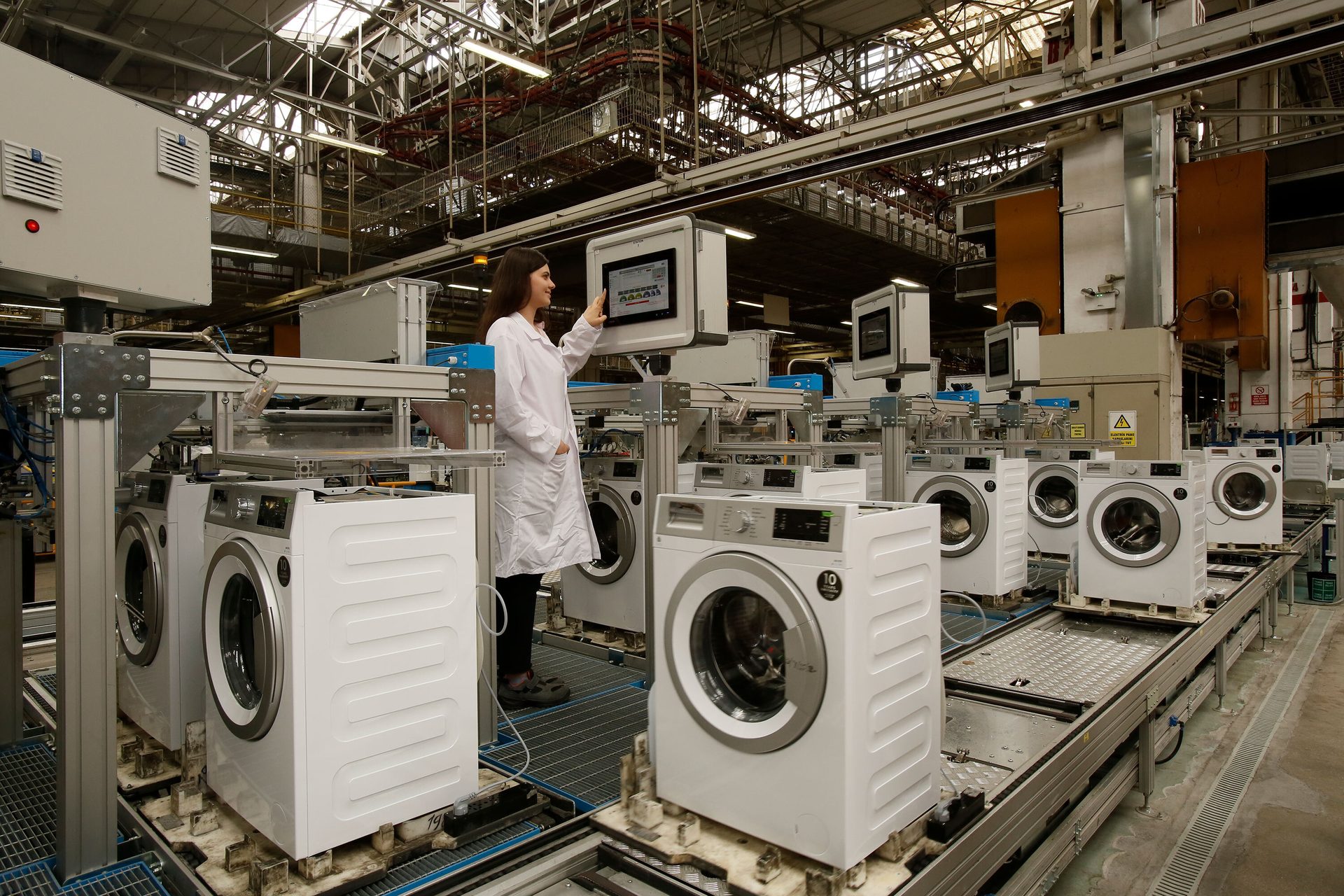 Woman in lab coat checking screen on washing machine factory line.