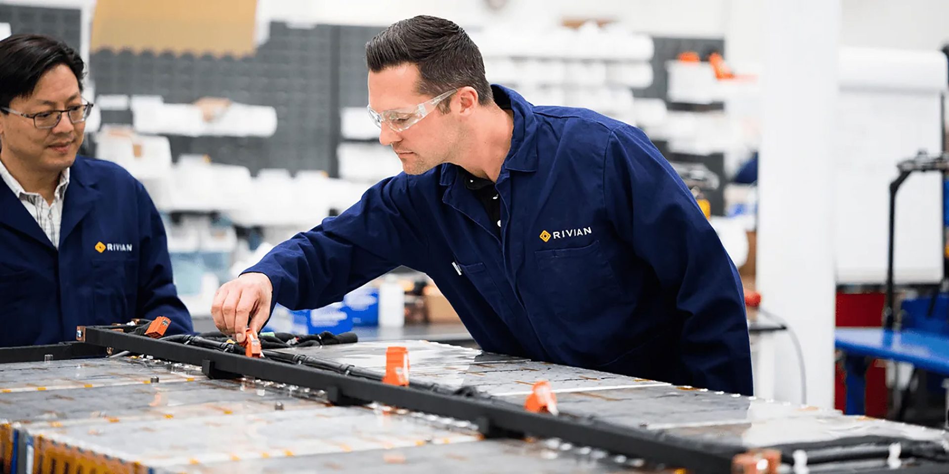 Two Rivian engineers in blue overalls inspect and work on an EV battery pack in a lab.