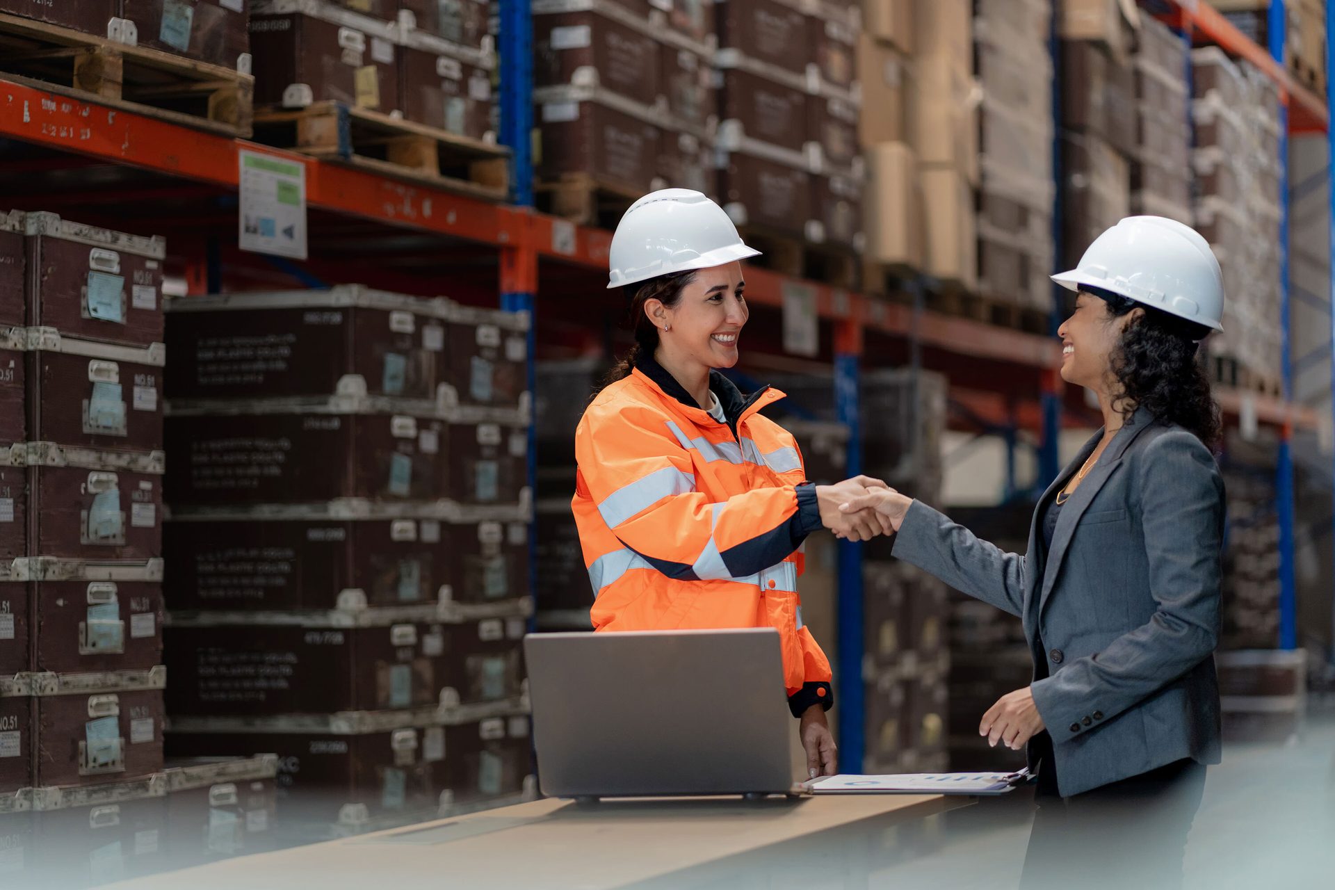 Two women in hard hats shake hands in a warehouse, one in safety gear, one in a suit.