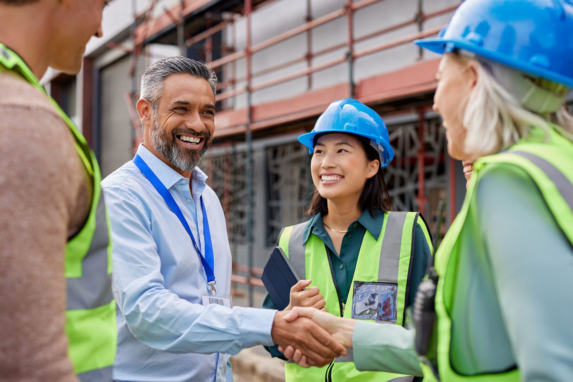 Four smiling construction workers in safety gear shake hands on site.
