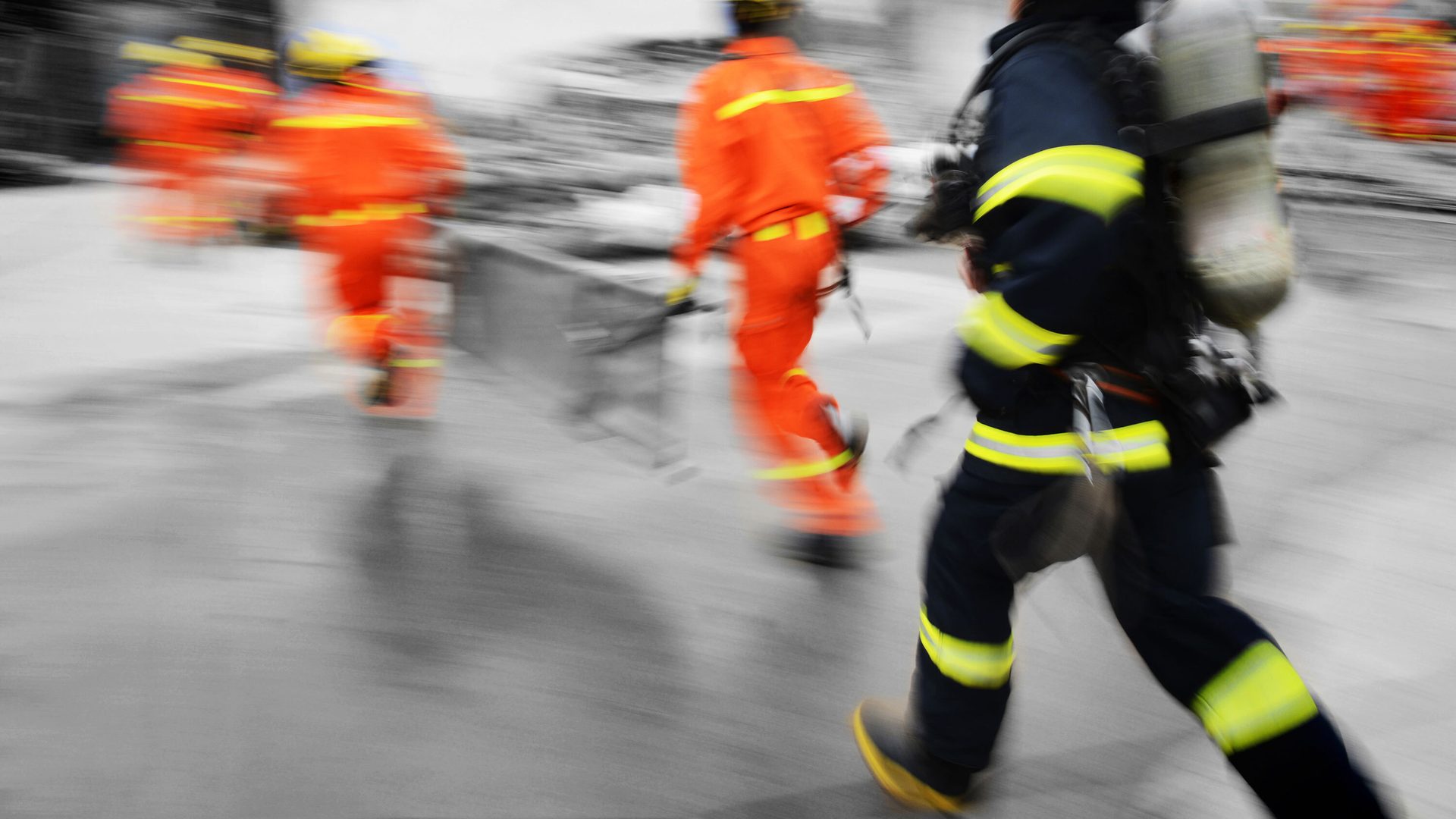 Motion-blurred firefighters in orange and blue gear, one with an oxygen tank, running to an emergency.