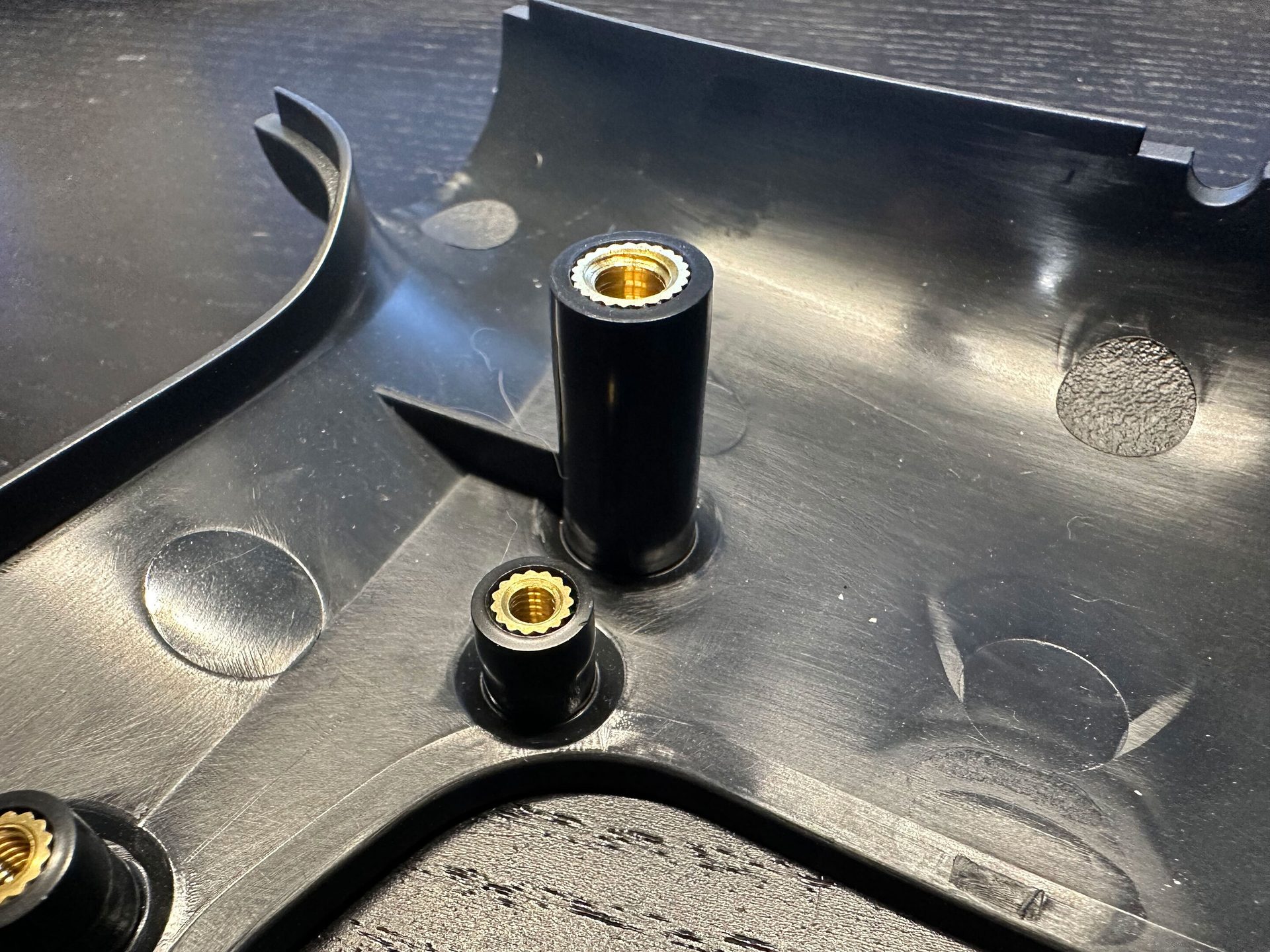 Black plastic part with two brass threaded inserts, one tall and one short, on a dark surface.