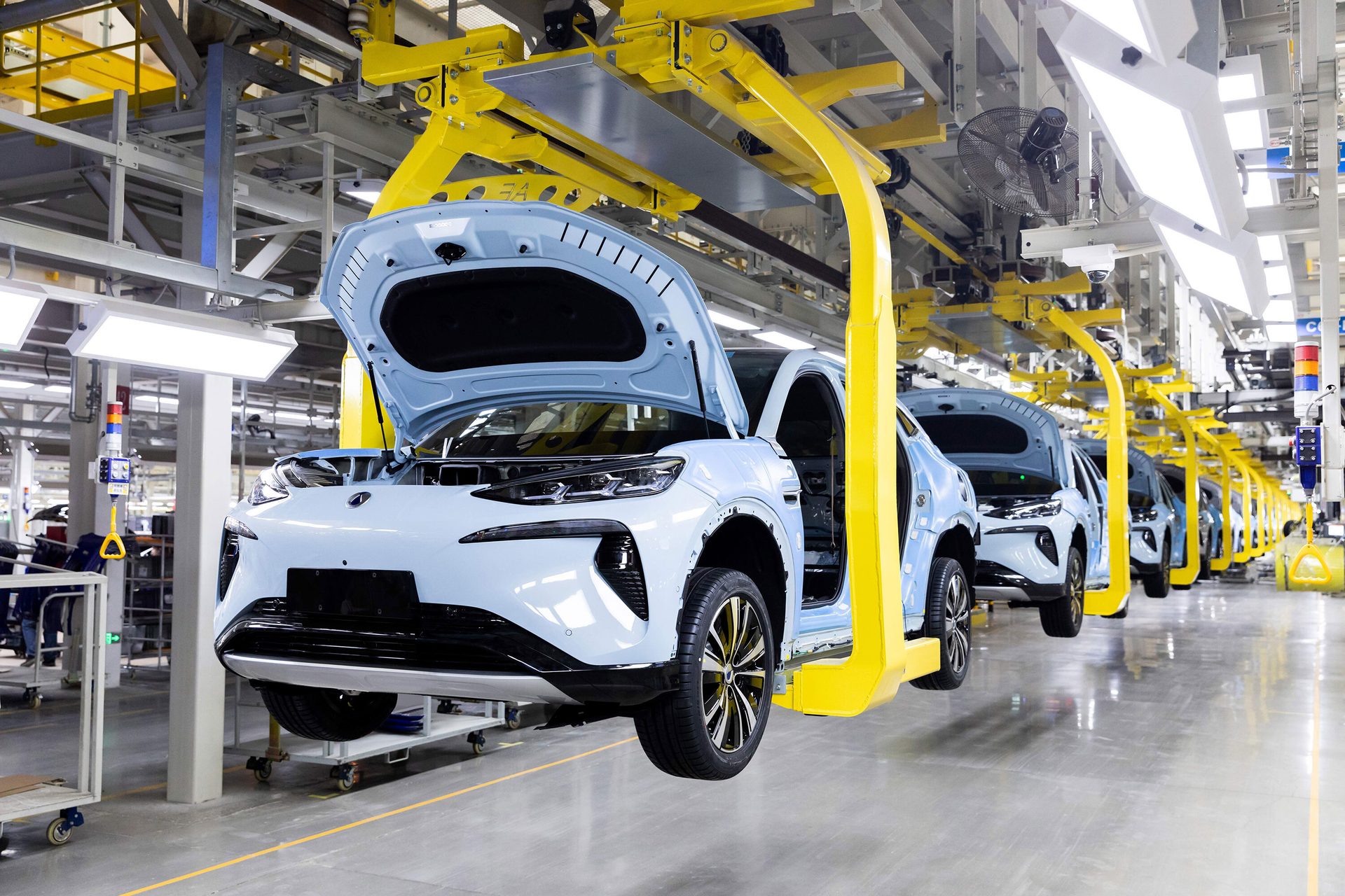 Light blue cars with open hoods move along an assembly line on a yellow overhead conveyor system in a factory.