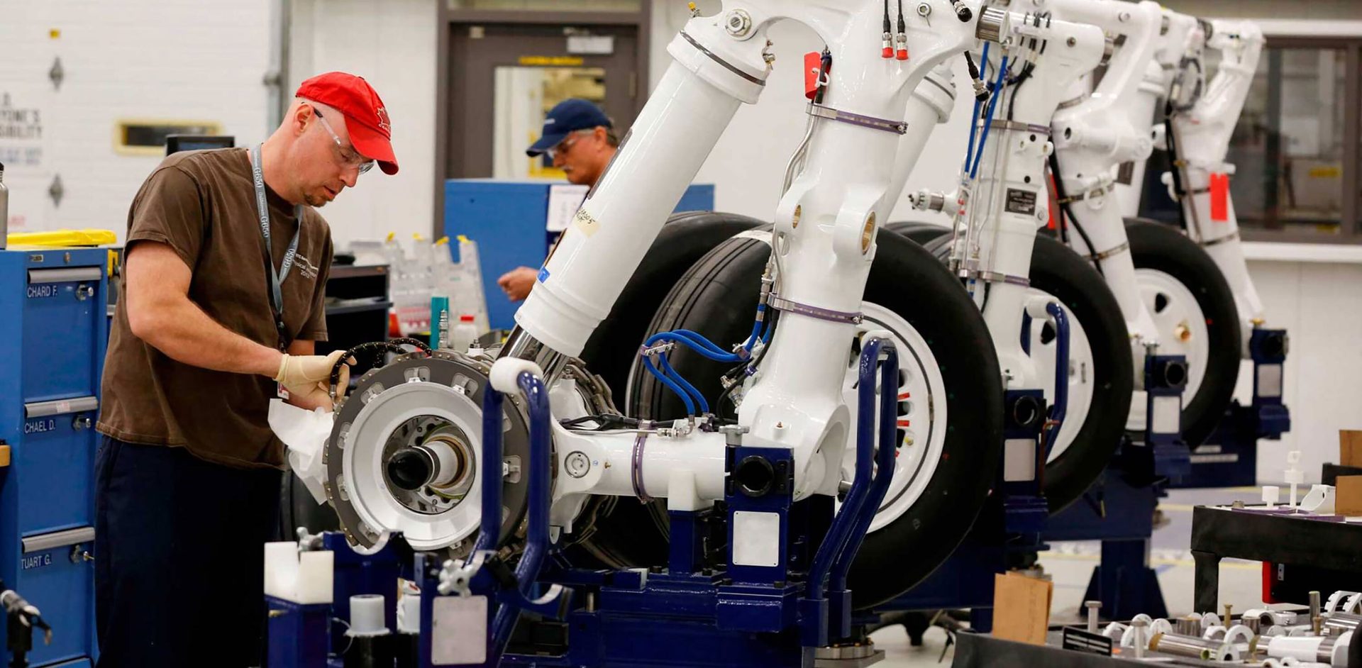 A technician assembles aircraft landing gear components while another works in the background.