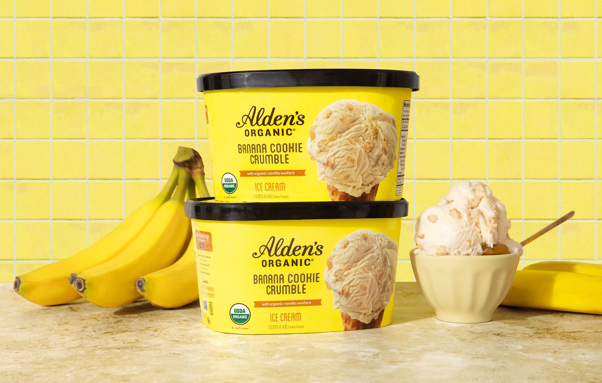 Two tubs of Alden's Organic Banana Cookie Crumble ice cream with bananas and a scoop in a bowl.