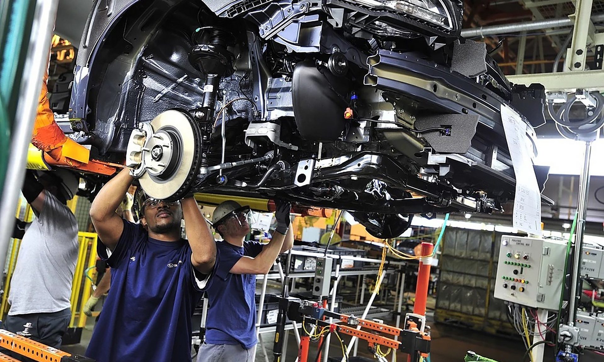 Two factory workers on an assembly line attach a brake disc to the underbody of a car during production.
