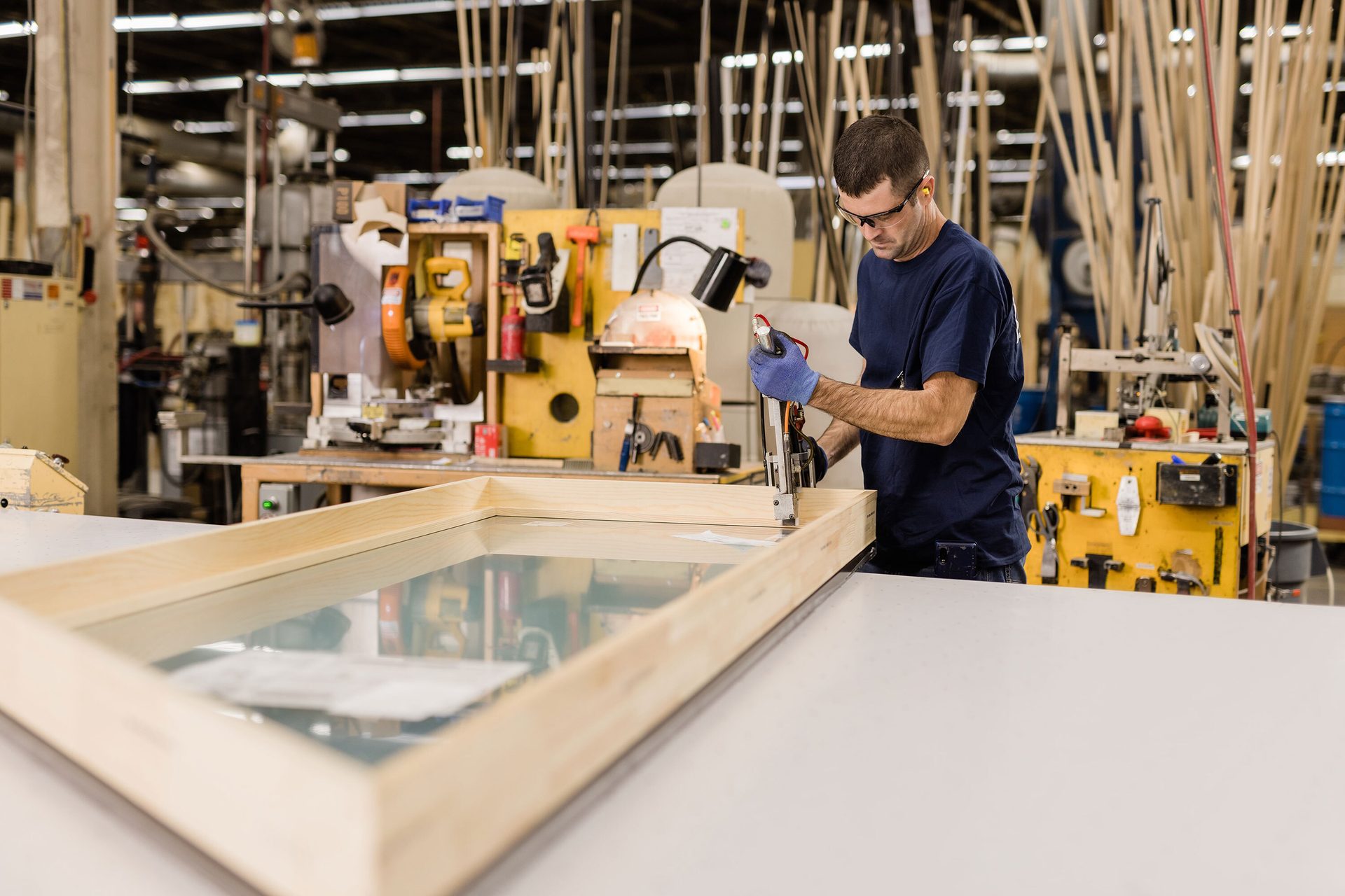 Man in safety glasses and gloves uses a tool on a wooden window frame with glass in a workshop.