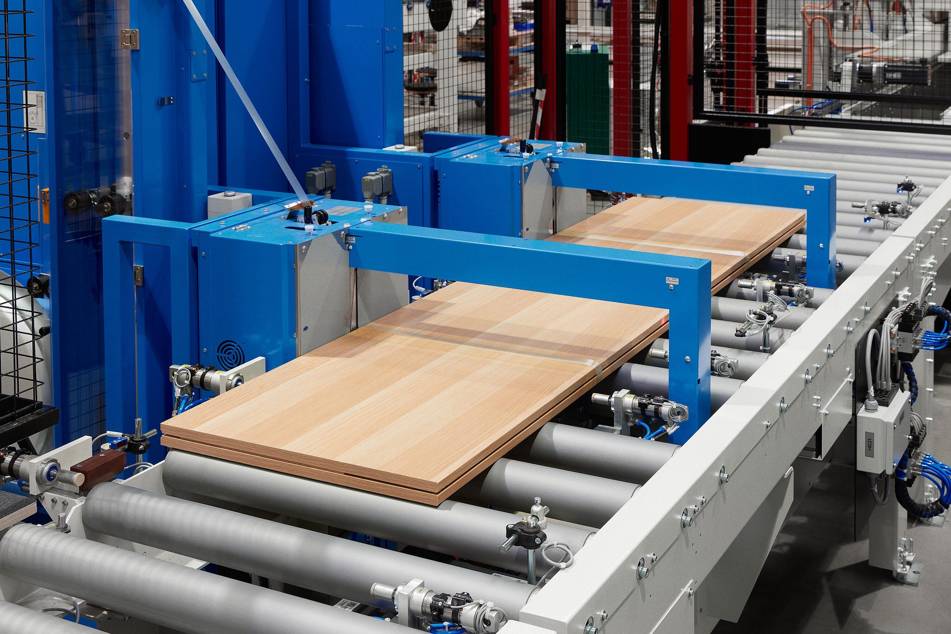 Wooden panels move along a roller conveyor system with blue industrial machinery for processing.